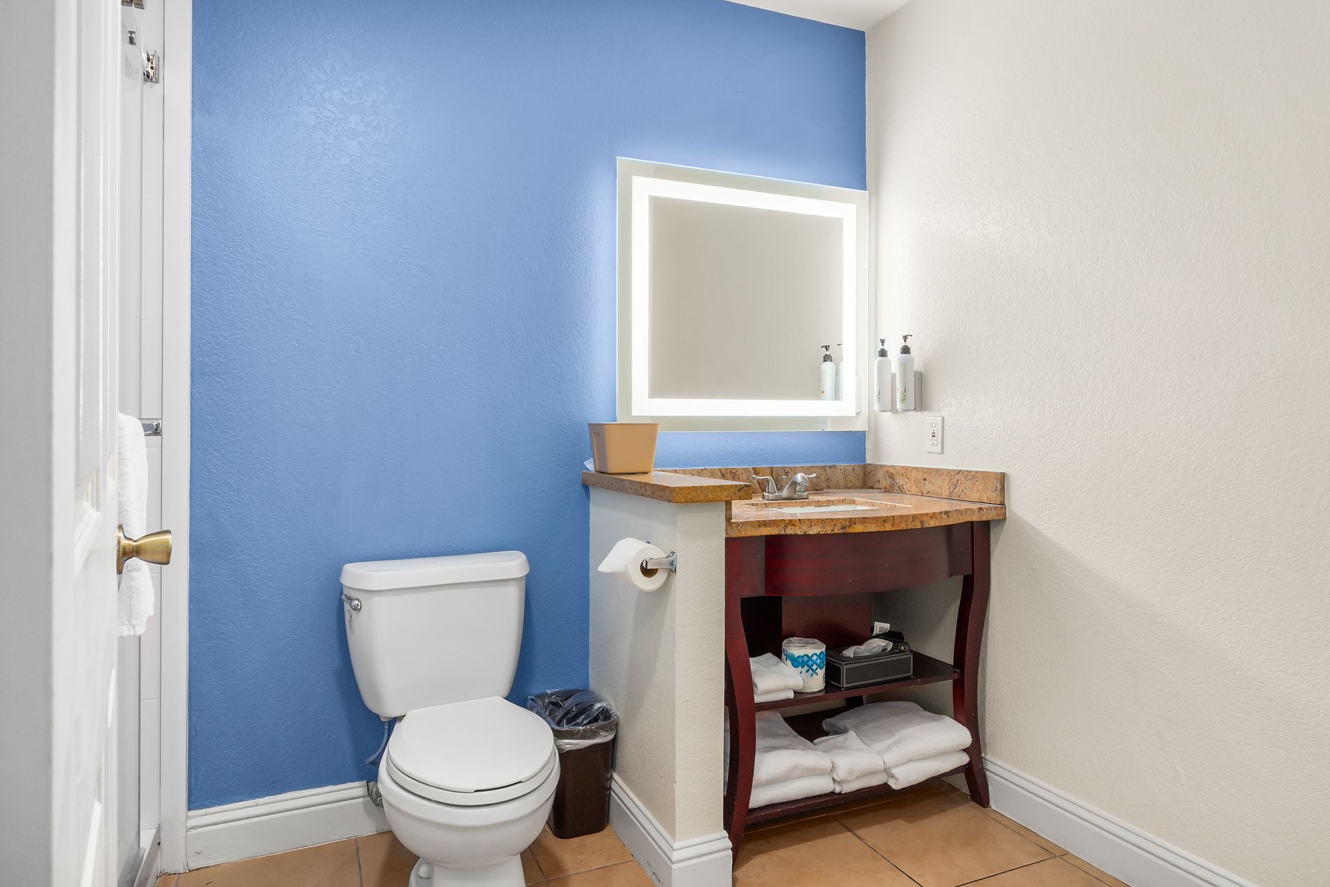 A bathroom with a blue wall, a white toilet, an illuminated square mirror, and a wood vanity with open shelving.