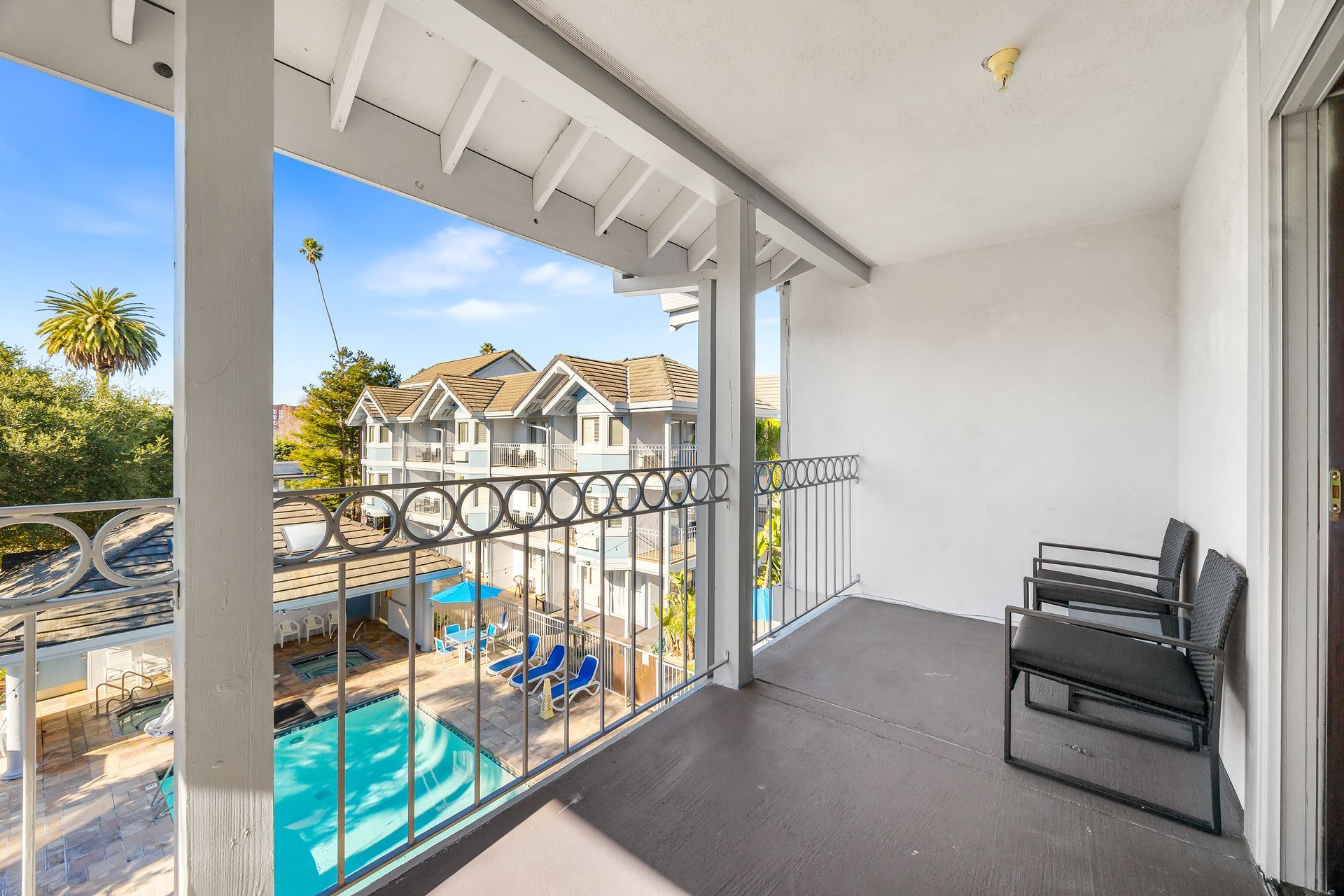 A balcony with two black chairs overlooks a swimming pool and neighboring apartment buildings under a clear blue sky.