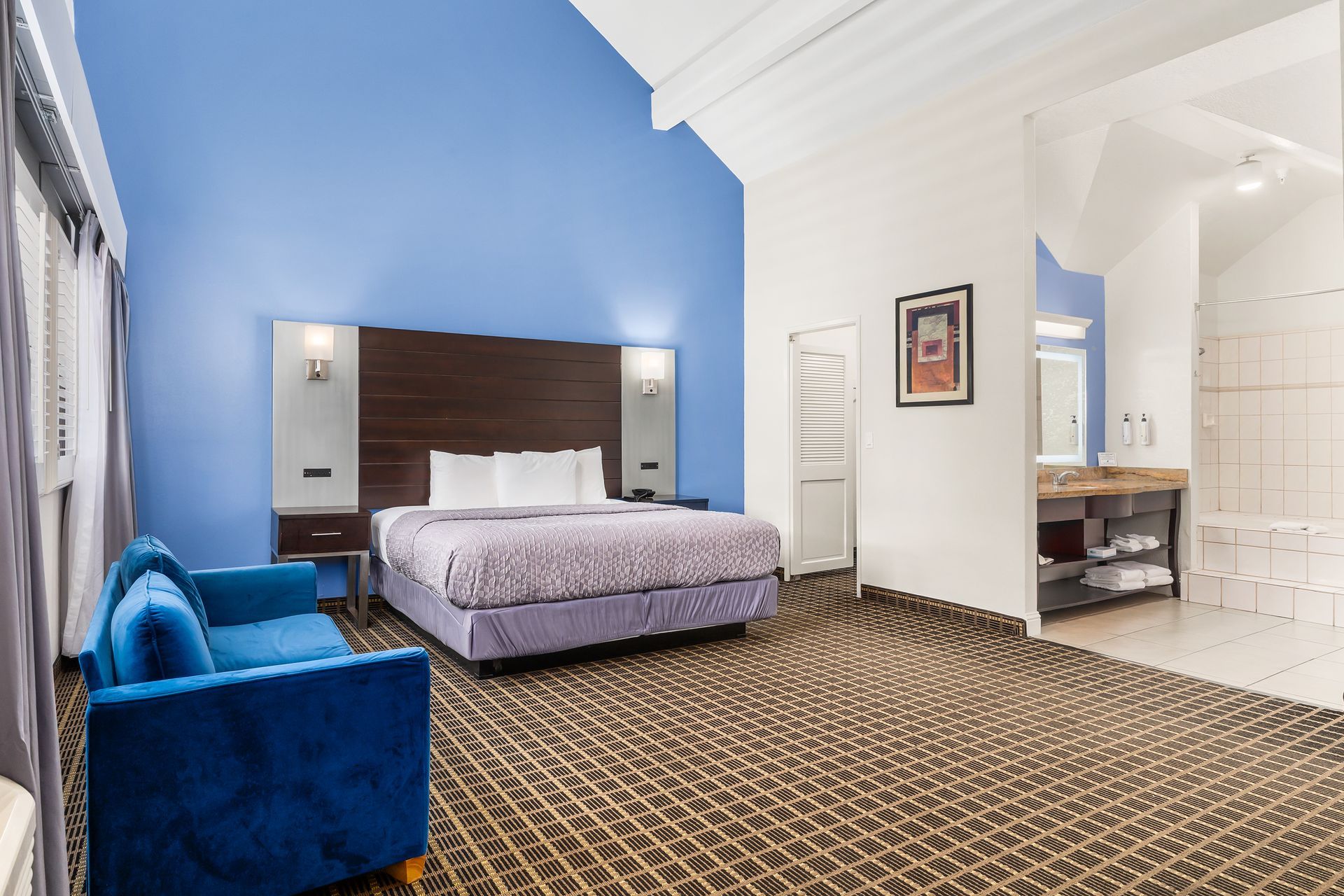 A spacious hotel room with a king bed, a blue sofa, brown patterned carpet, and an open, light-colored bathroom.