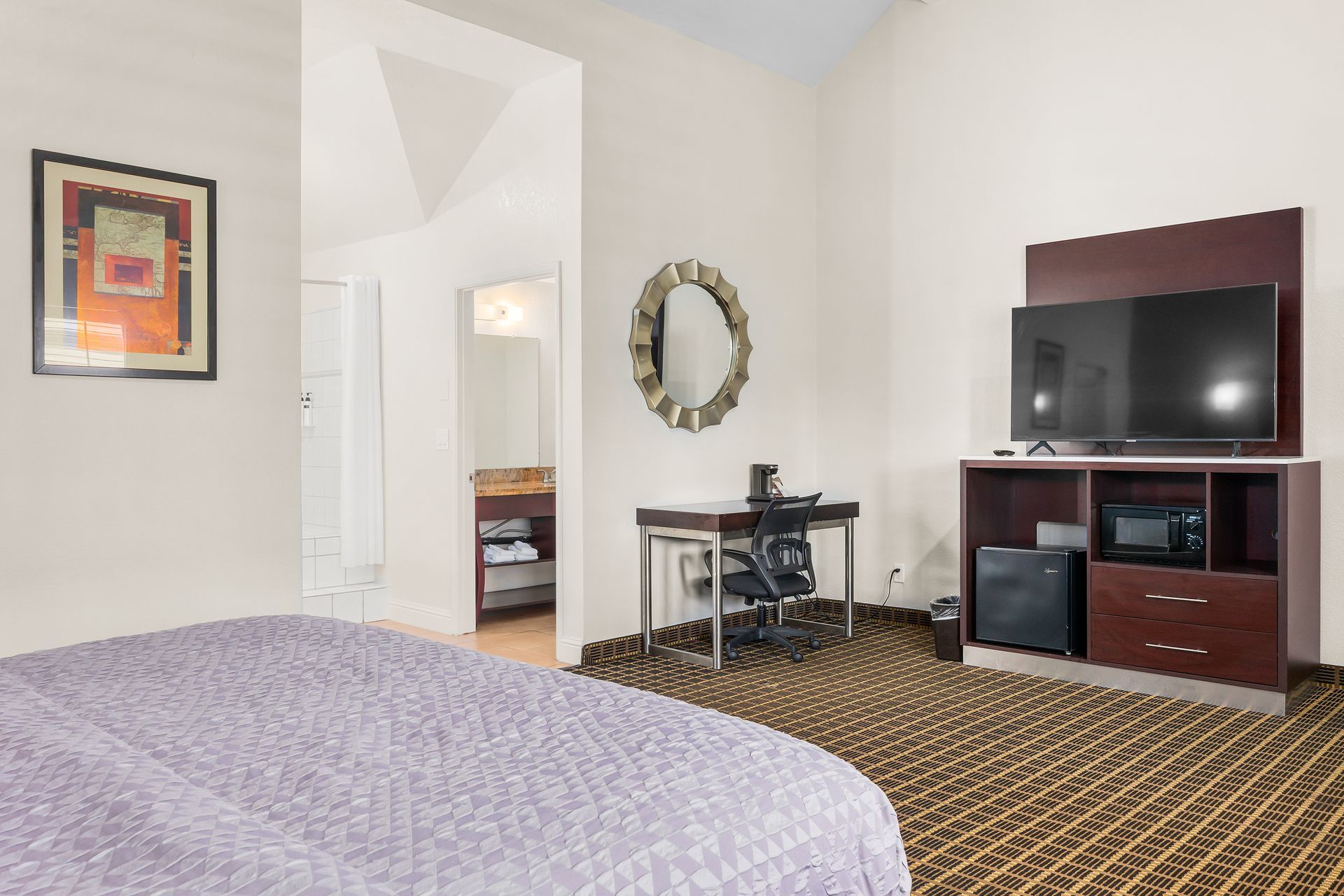 A hotel room with a lavender bed, a desk, a mirror, and a television on a wooden media console with a small refrigerator.