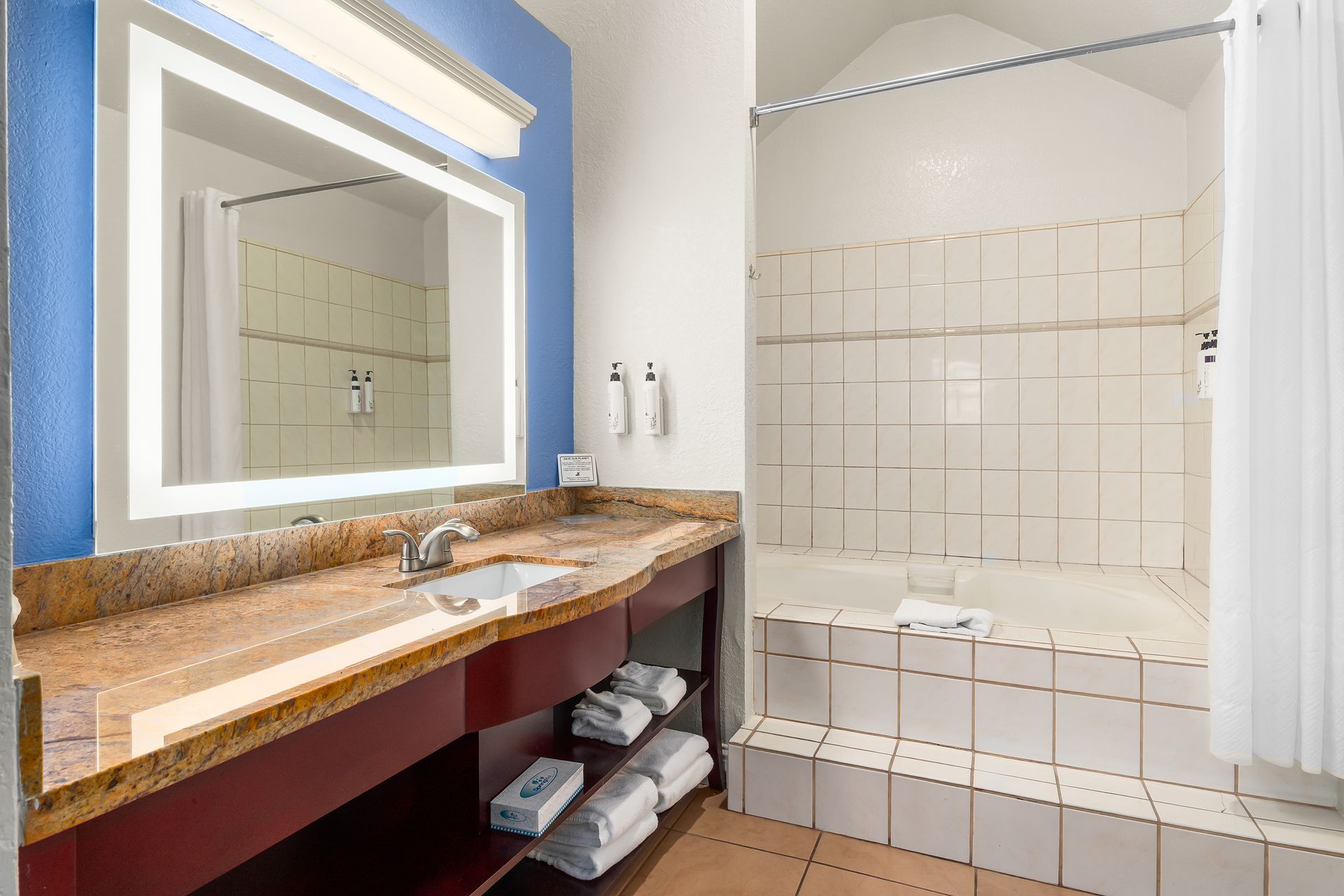 A bathroom featuring a granite vanity, an illuminated mirror, and a tiled bathtub with a shower curtain.