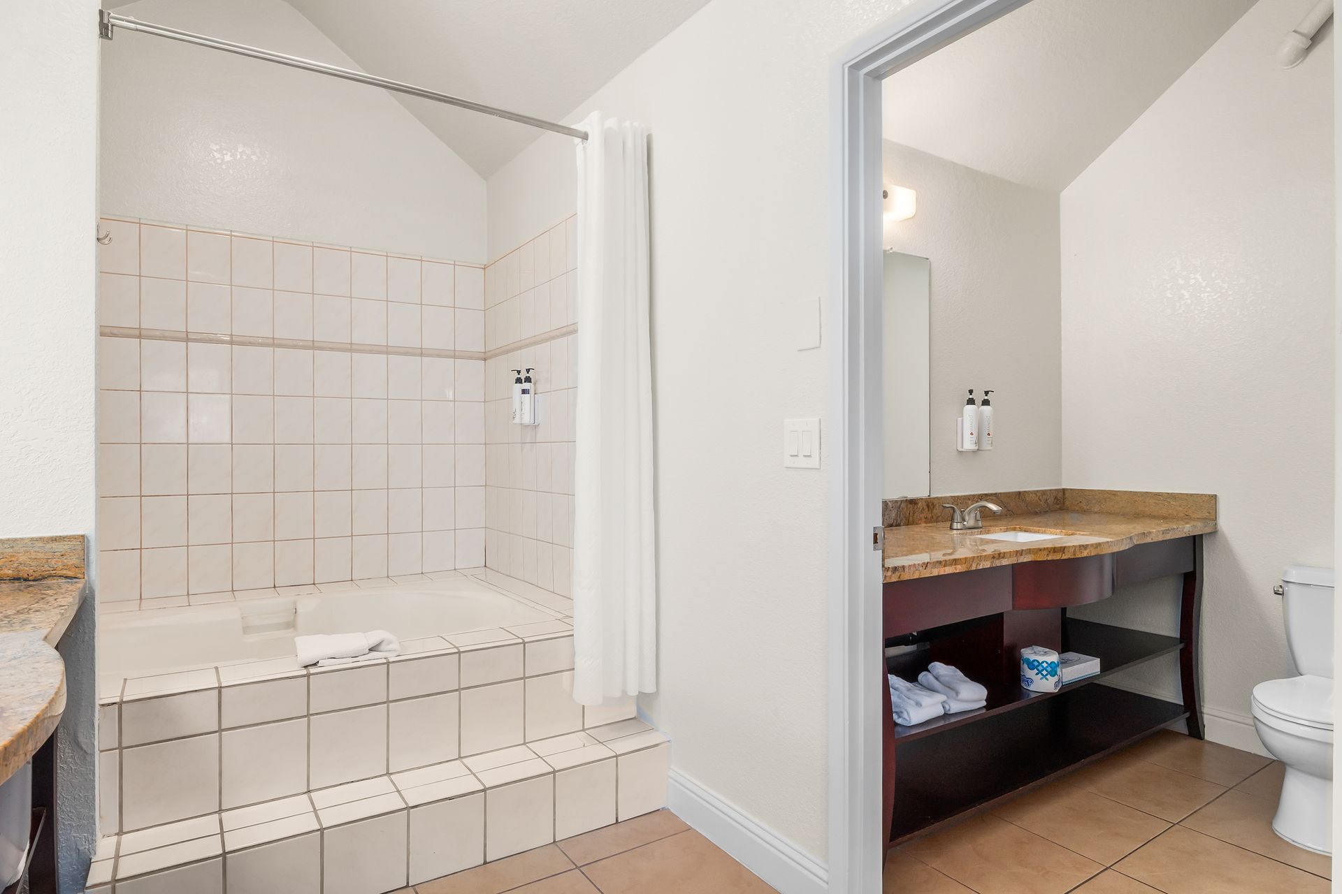 A brightly lit bathroom with a tiled bathtub area, a separate vanity with a mirror, and a toilet.
