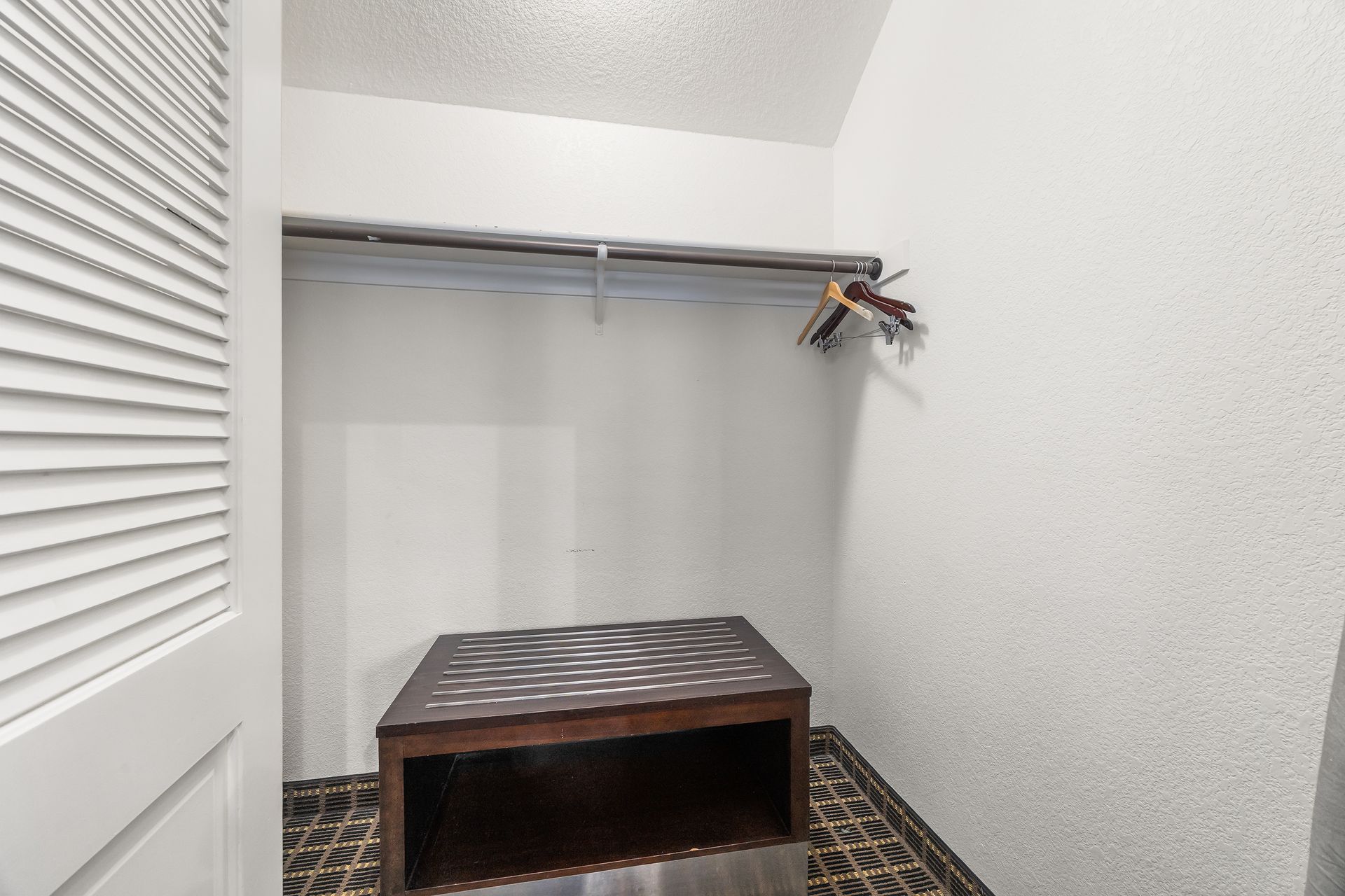 A small walk-in closet with white walls, a metal clothing rod, and a low wooden storage bench on a dark patterned floor.