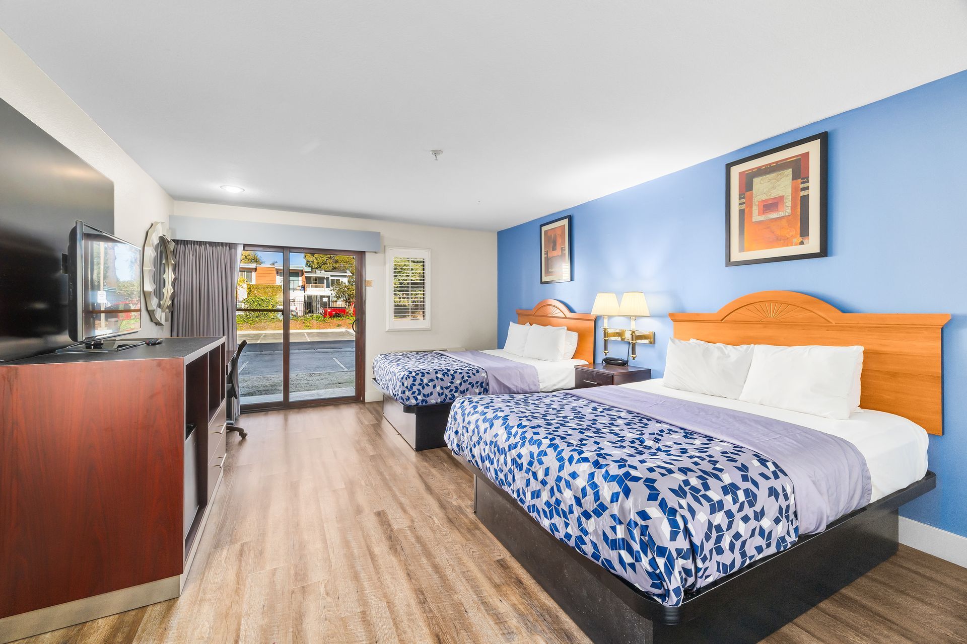 A hotel room with two beds, wood floors, a television, and a sliding glass door against a light blue wall.