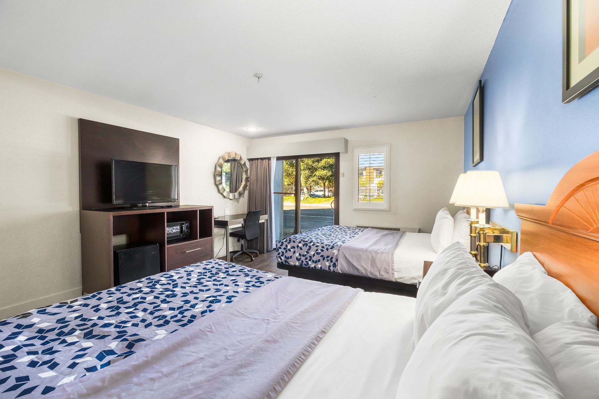 A brightly lit hotel room featuring two beds with patterned blue and white runners, a television stand, and a desk.