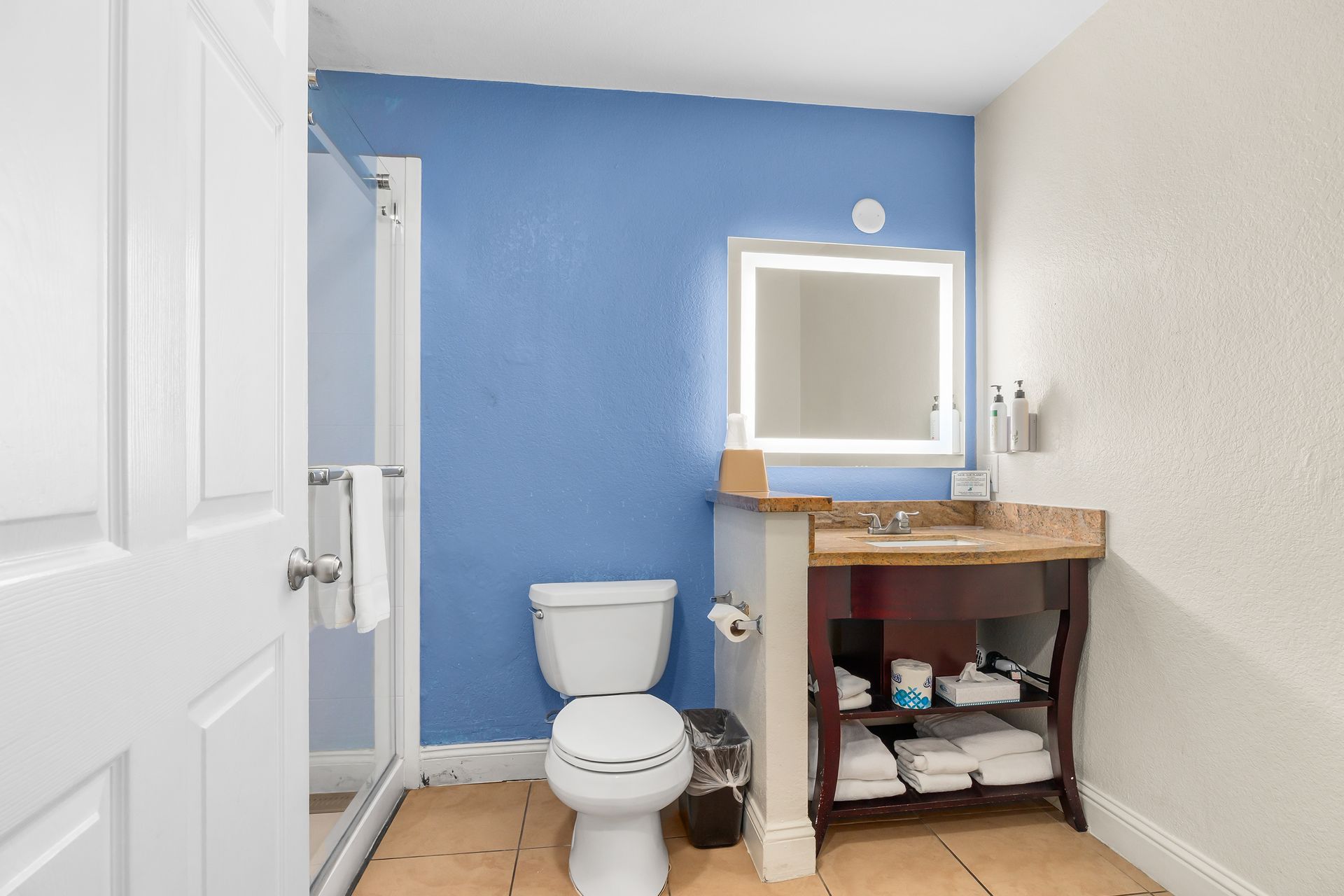 A bathroom with a blue wall, white toilet, a lighted vanity mirror, and an open storage shelf with towels under the sink.
