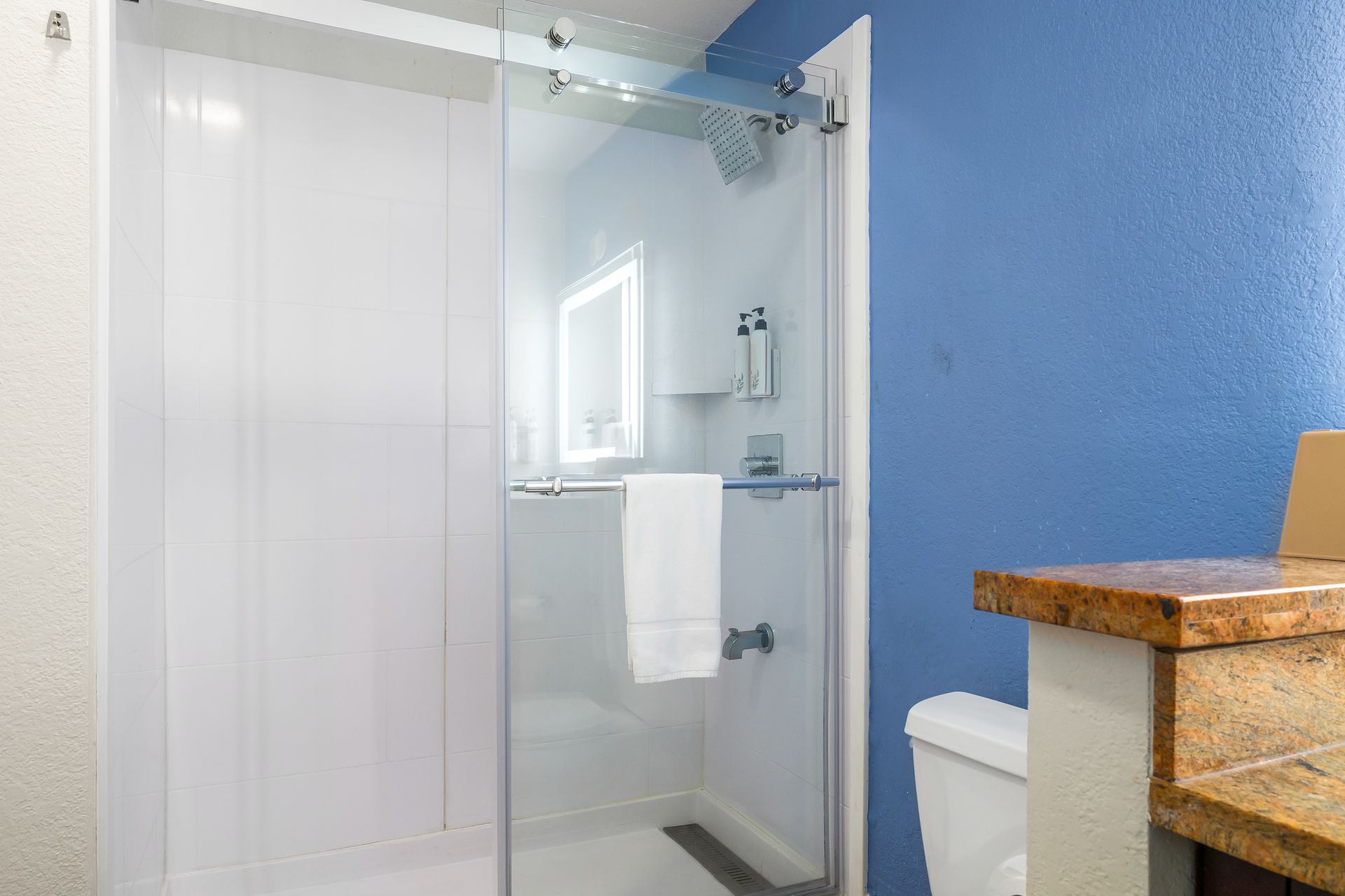 A glass-enclosed walk-in shower with a white towel on the handle, next to a blue wall and a granite countertop.
