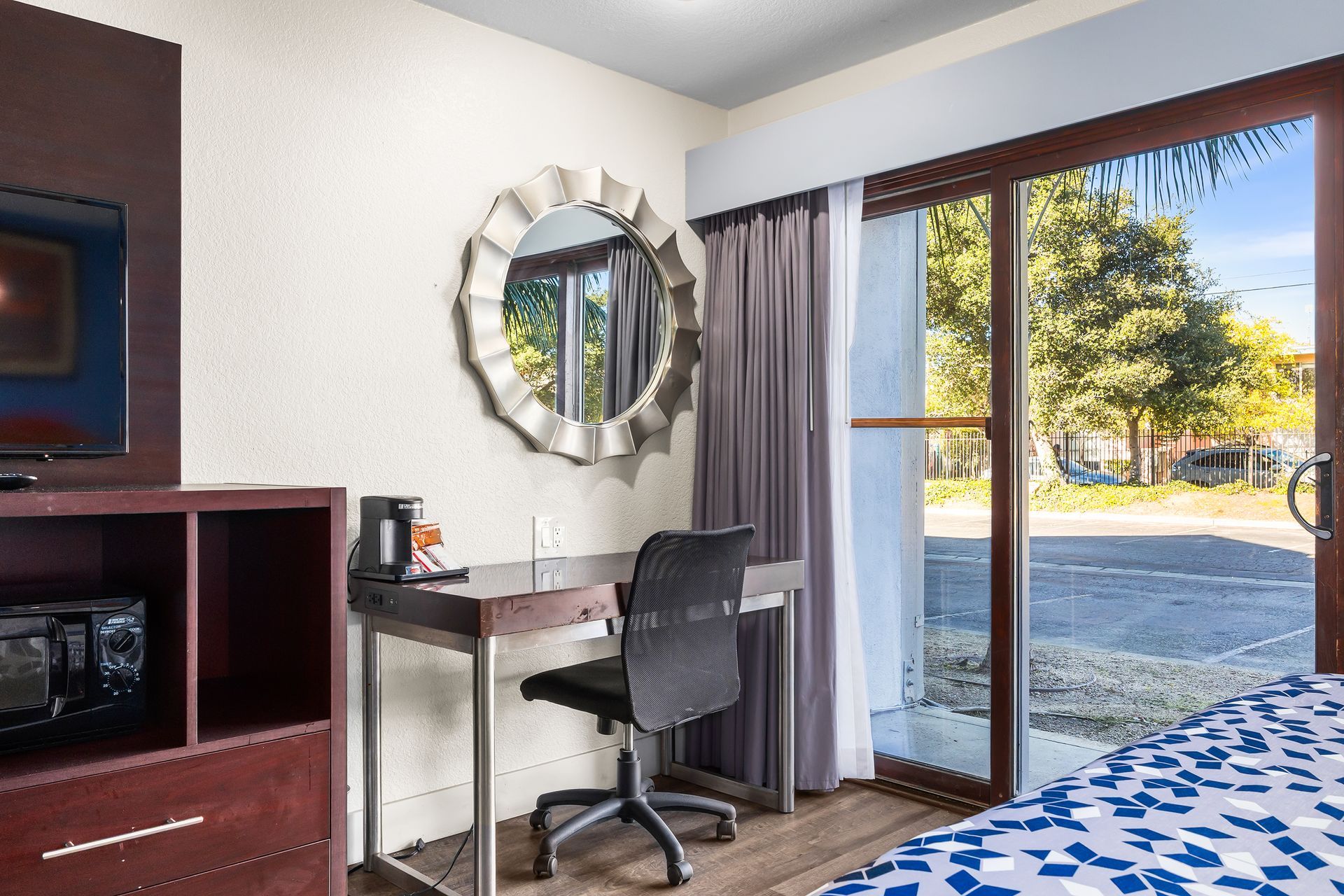 A hotel room corner with a desk, office chair, round mirror, TV stand, and sliding glass door to an outdoor area.