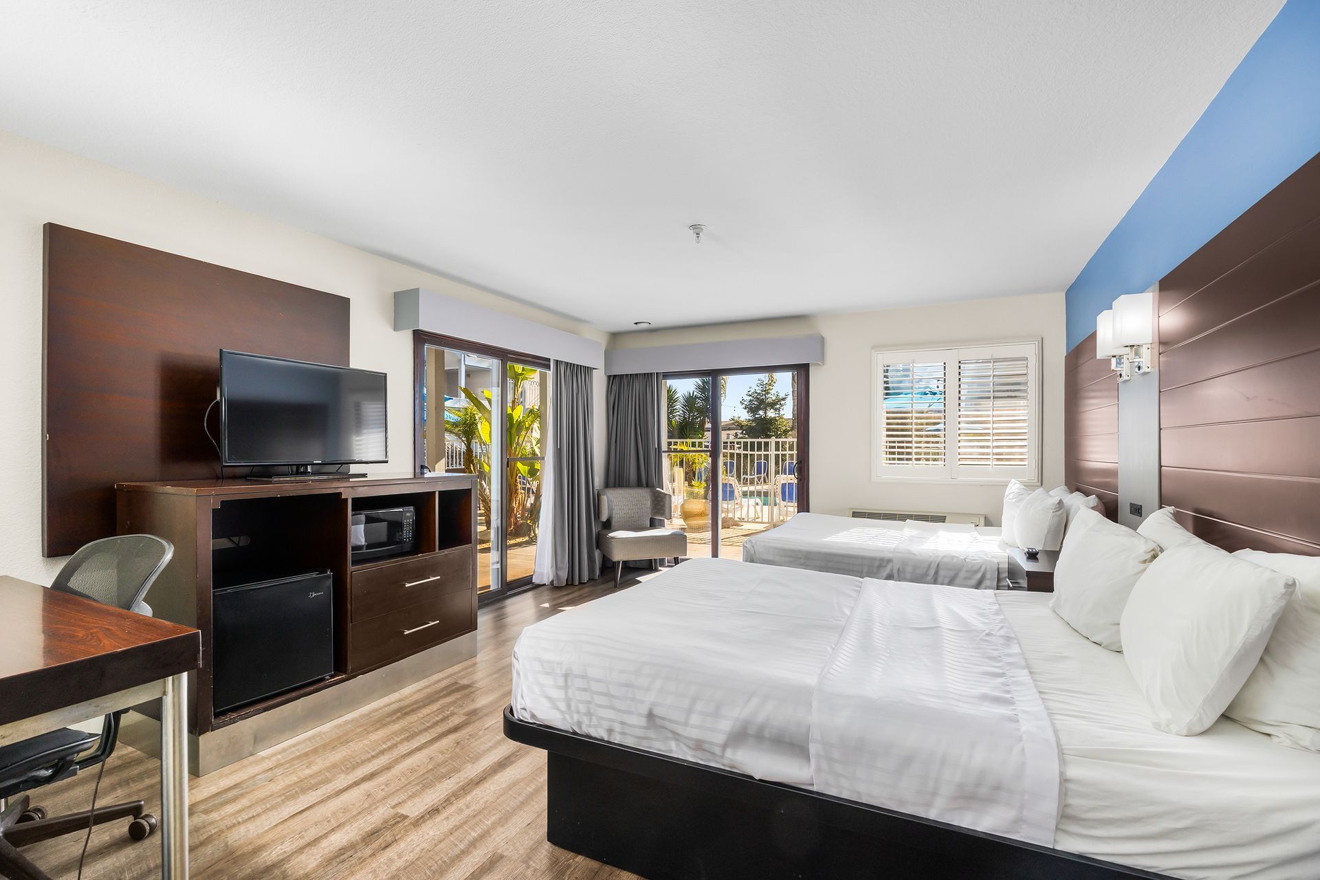 A modern hotel room with two beds, a desk, a TV console, a small armchair, and a sliding door leading to a balcony.