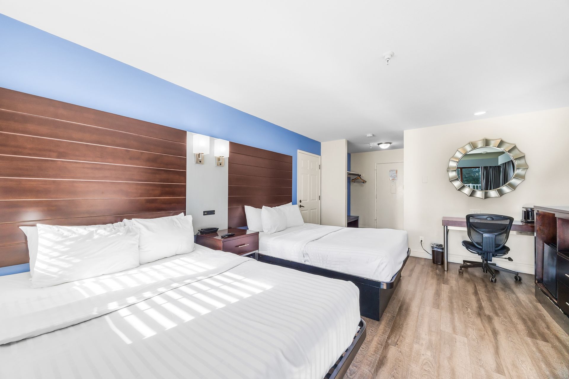 A hotel room with two white-bedded queens, a wooden headboard, light blue accent wall, desk, and round silver mirror.