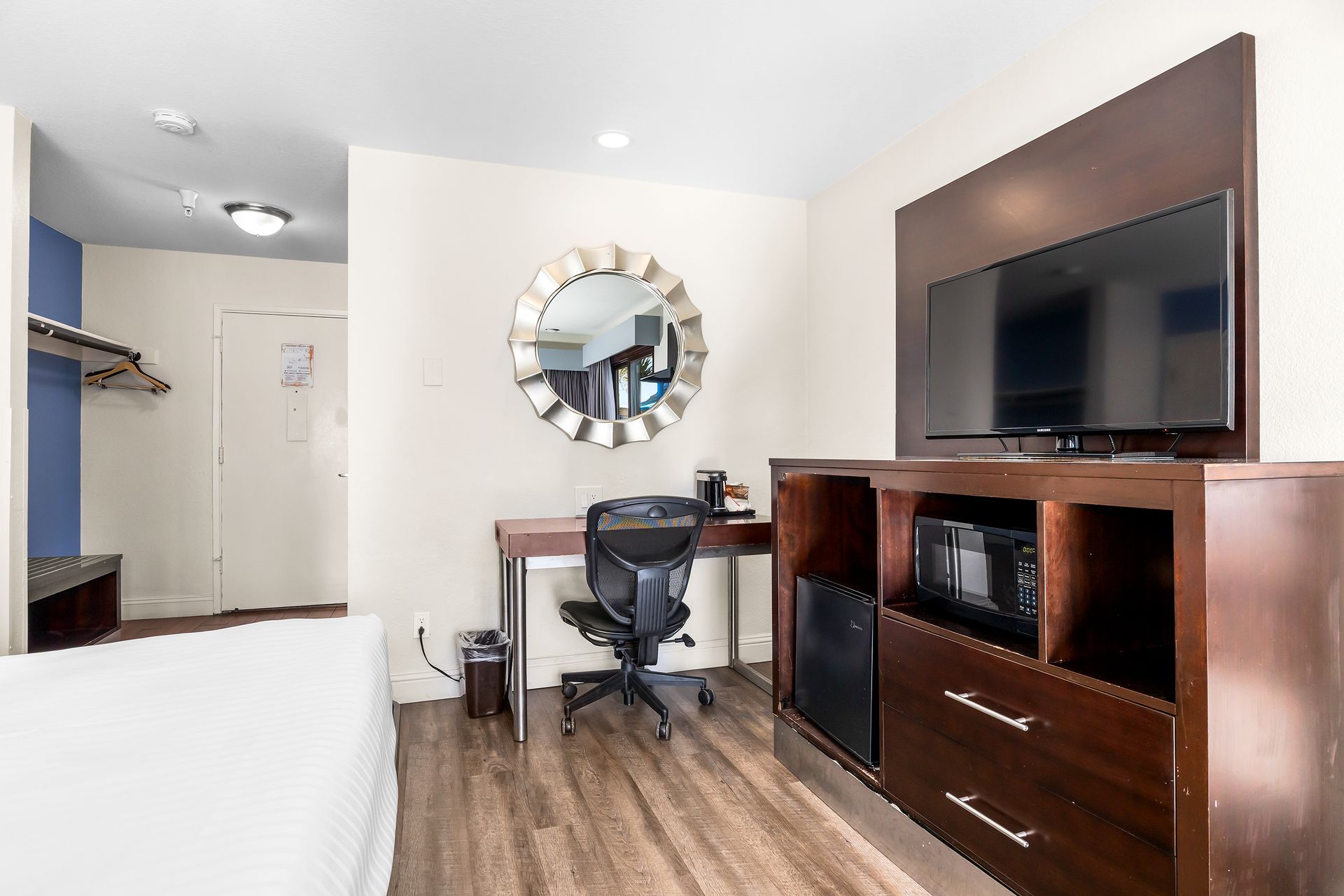 A hotel room interior featuring a desk with a rolling chair, a circular mirror, and a television on a dark wood dresser.