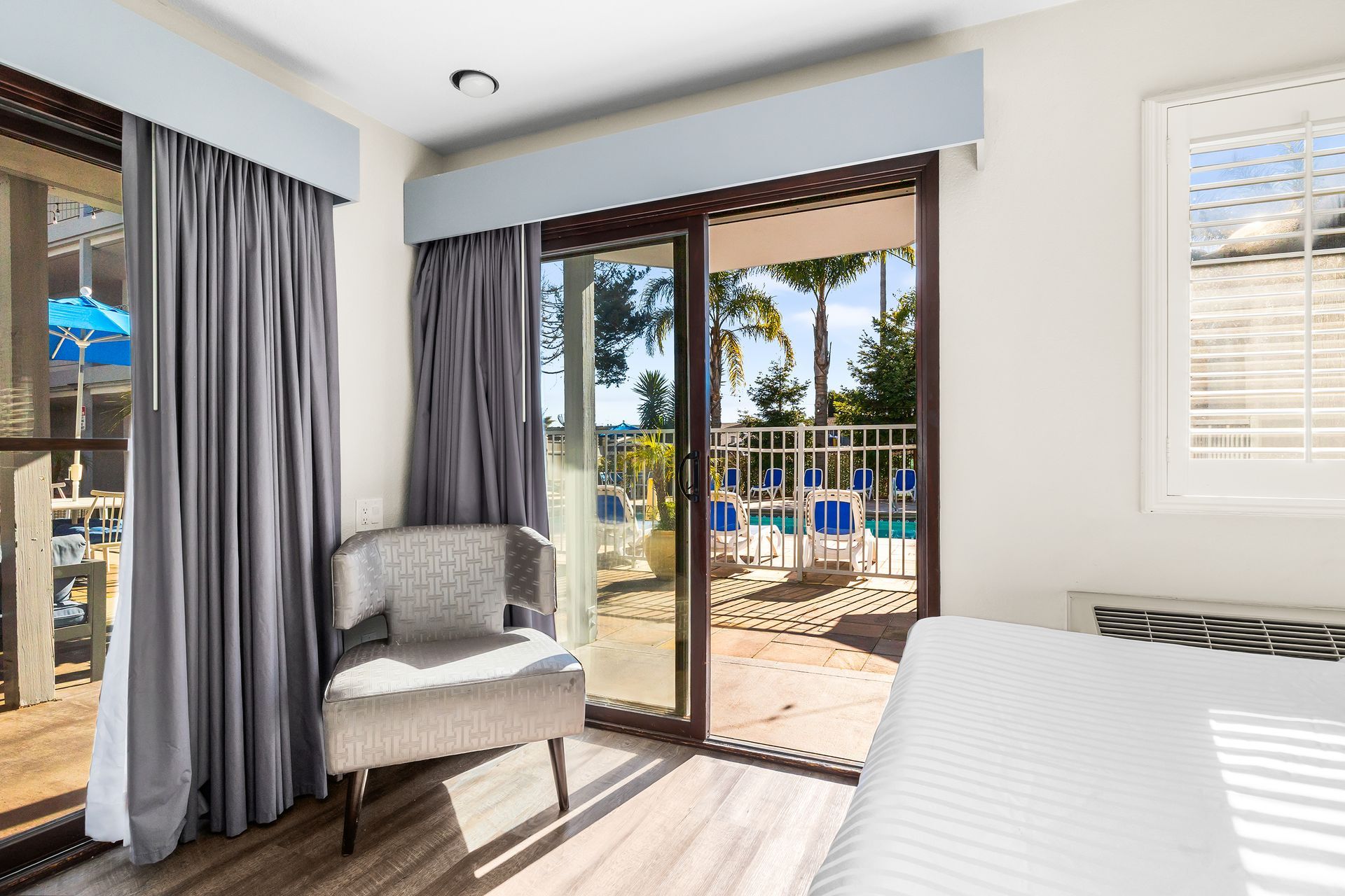 A hotel room with a gray armchair, floor-to-ceiling curtains, and a sliding glass door opening to a pool patio.