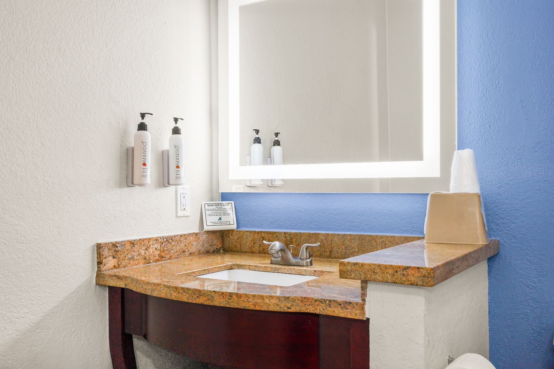 A bathroom vanity with a tan stone countertop, dark wooden cabinet, illuminated mirror, and blue wall accents.