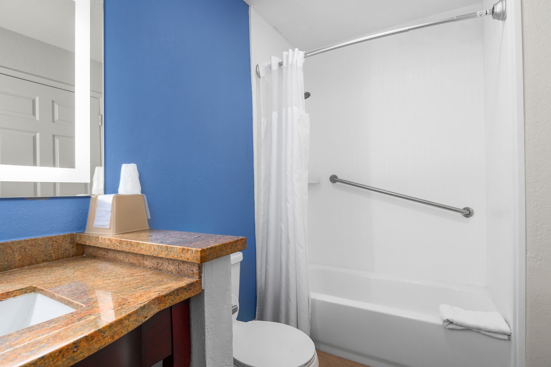 A bathroom with a blue wall, tan granite vanity countertop, white sink, and a bathtub with a shower curtain and grab bar.