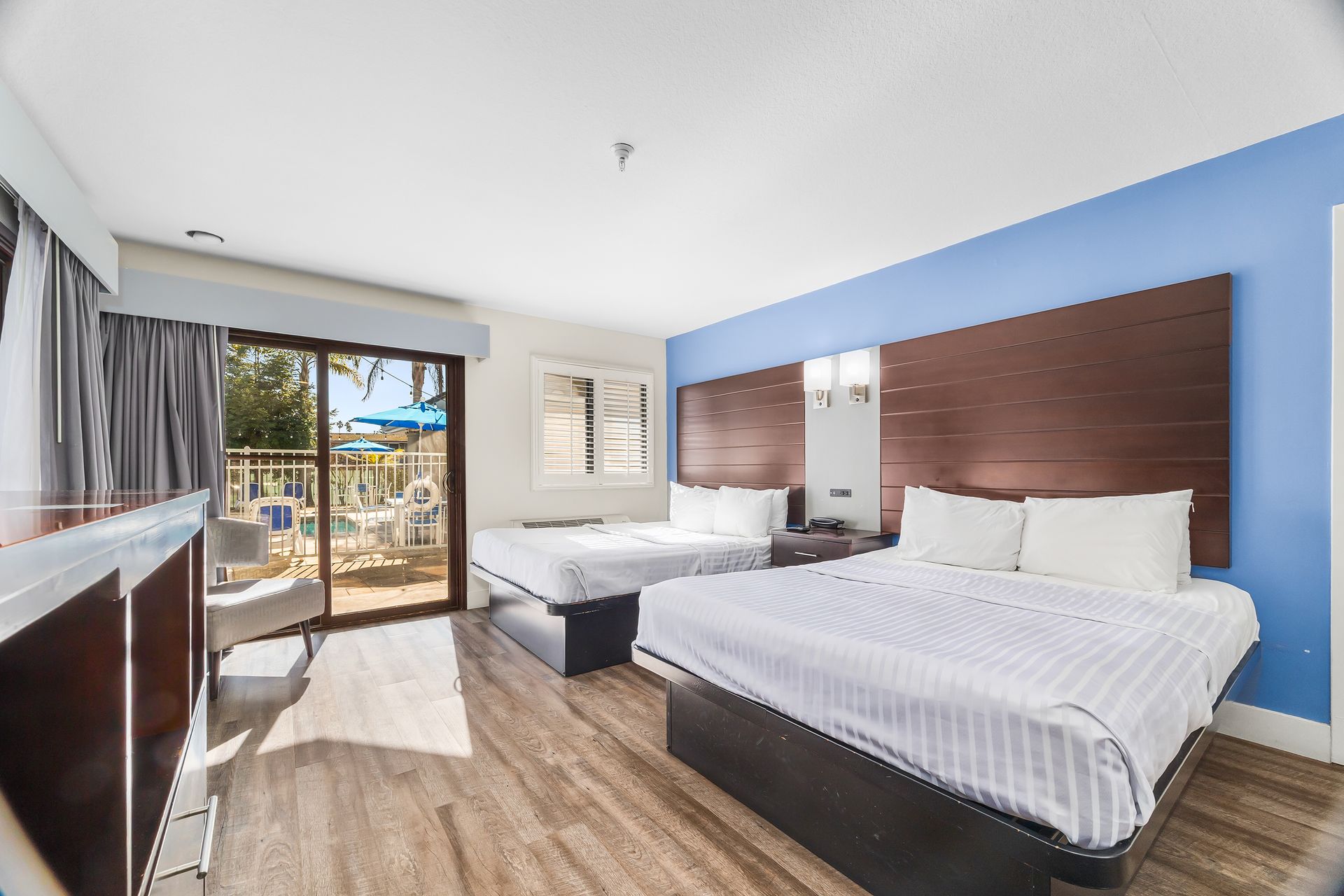 A hotel room with two white-bedded queens, dark headboards, wood floors, and a balcony view of a pool area.