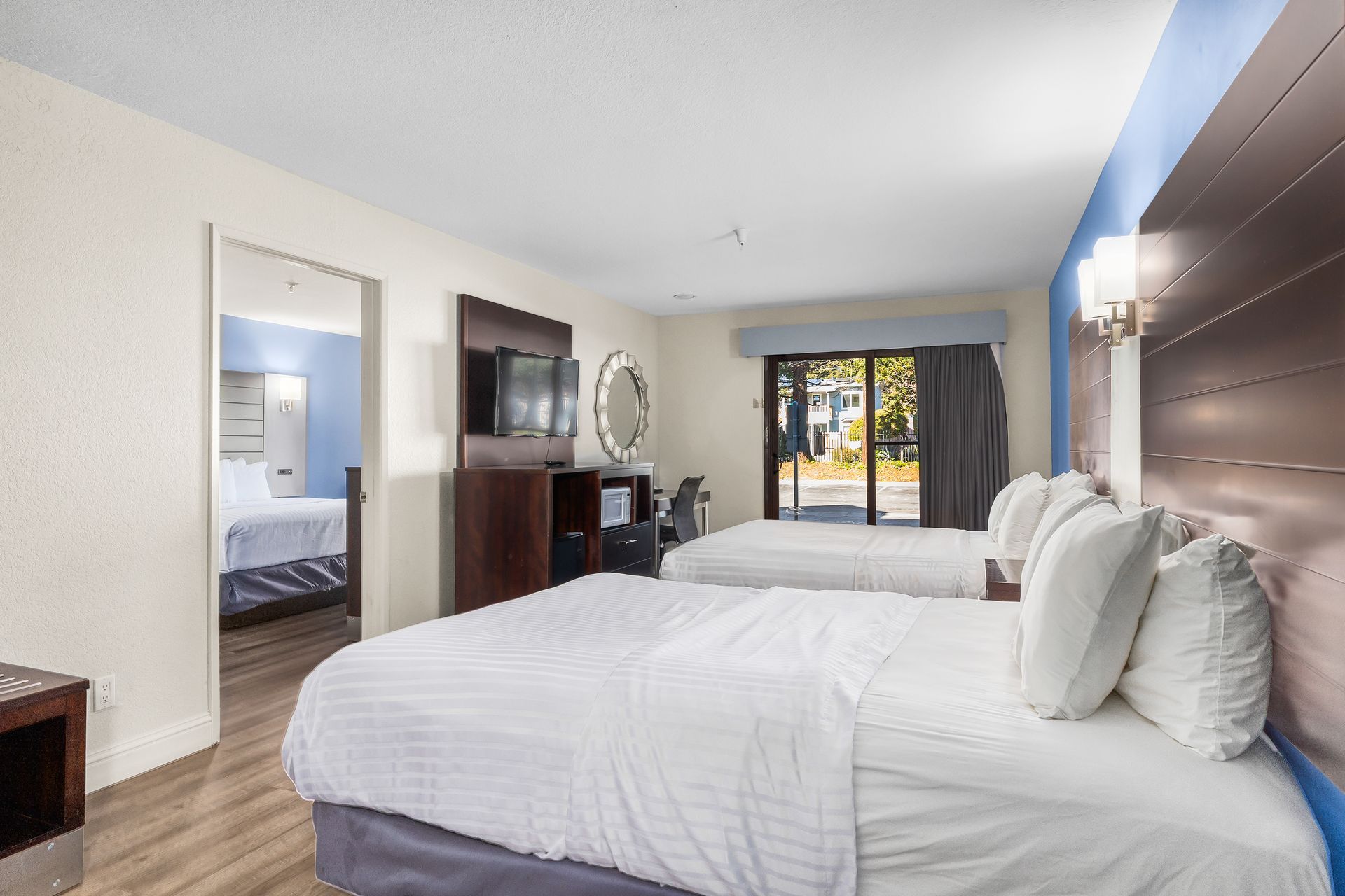 A hotel room with two white beds, a television on a dark wood stand, and an open door showing another room.