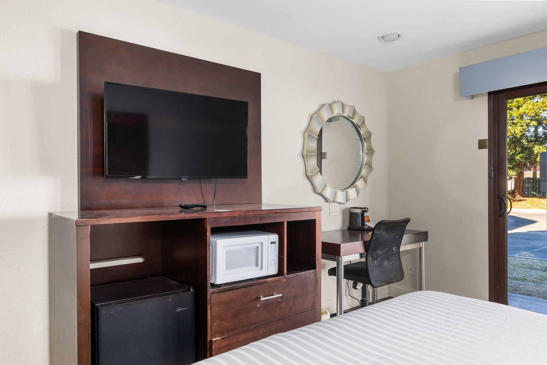 A hotel room featuring a wall-mounted TV, a dresser with a microwave and mini-fridge, a desk, and a sliding glass door.