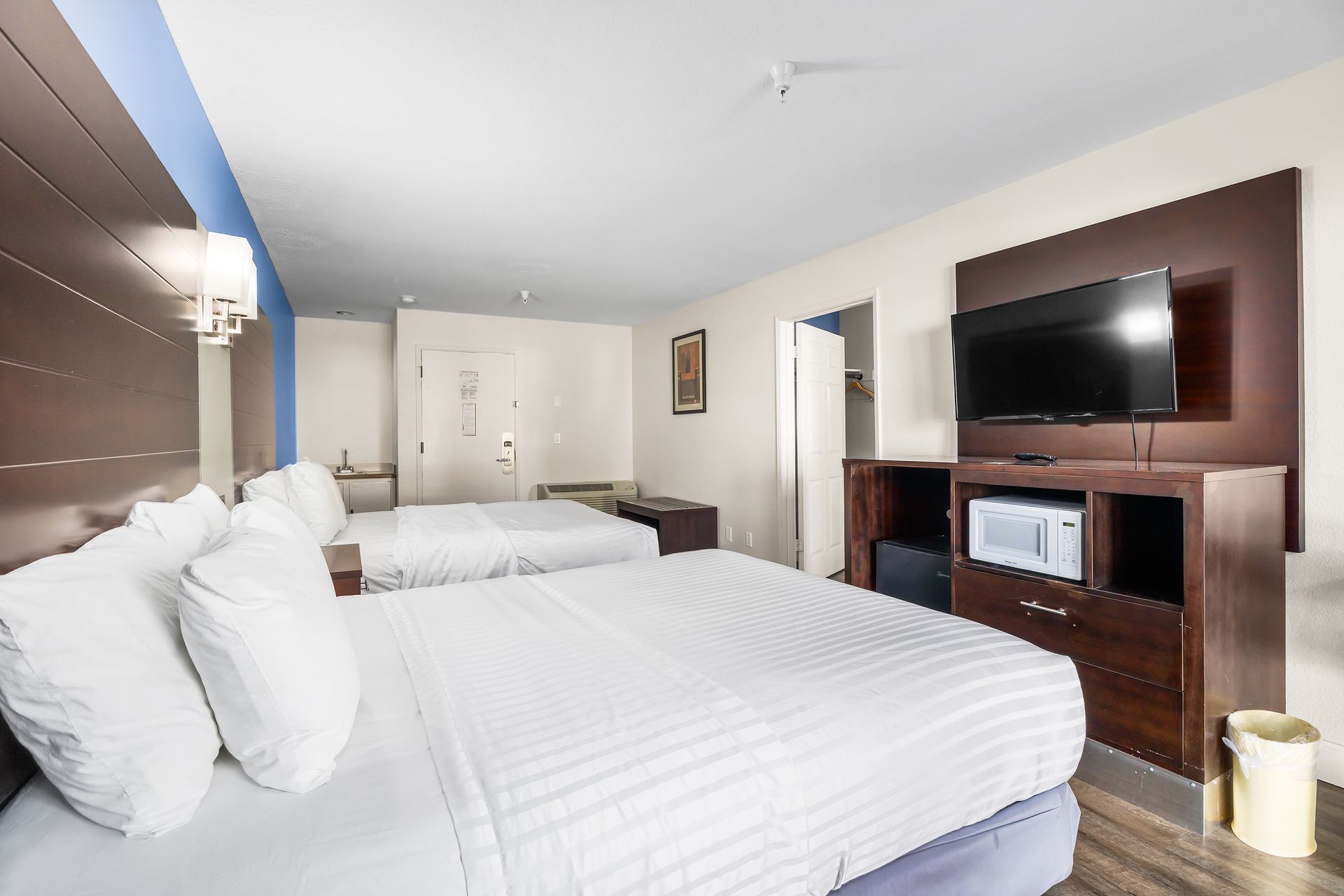 A hotel room featuring two queen beds with white linens, a dark wood dresser, a flat-screen television, and a trash bin.