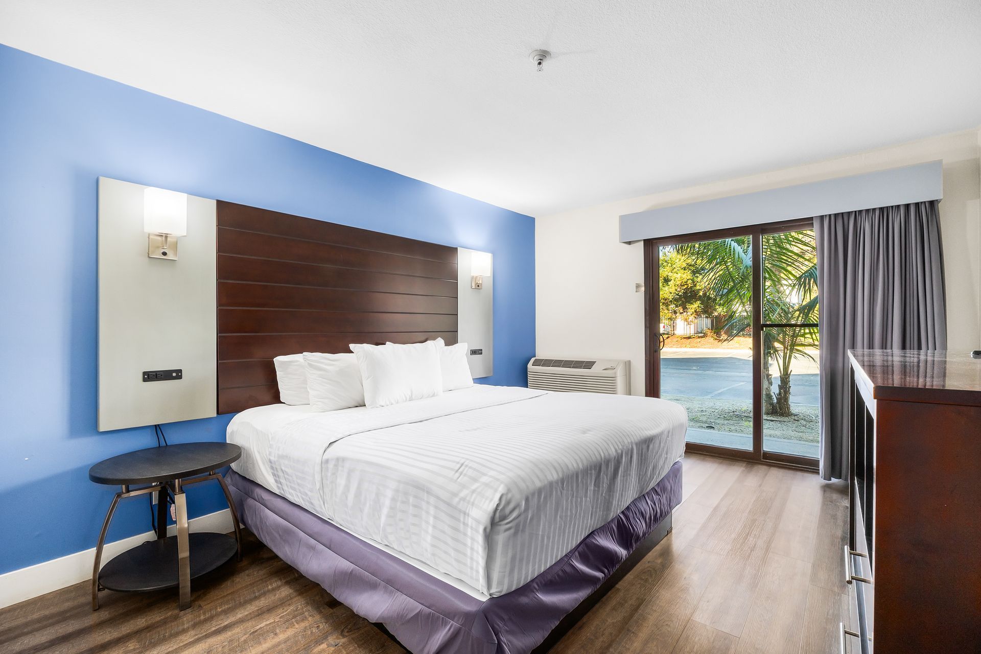 A hotel room featuring a king-size bed with white bedding, a wooden headboard, blue accent wall, and sliding glass doors.