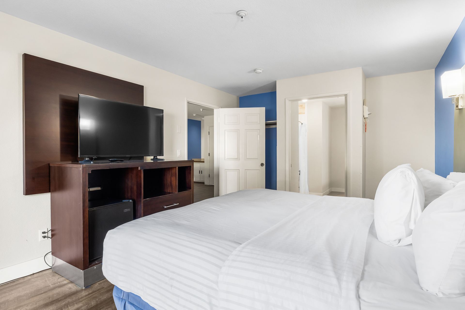 A hotel room features a large white bed with pillows, facing a dark wood media console topped with a flat-screen TV.