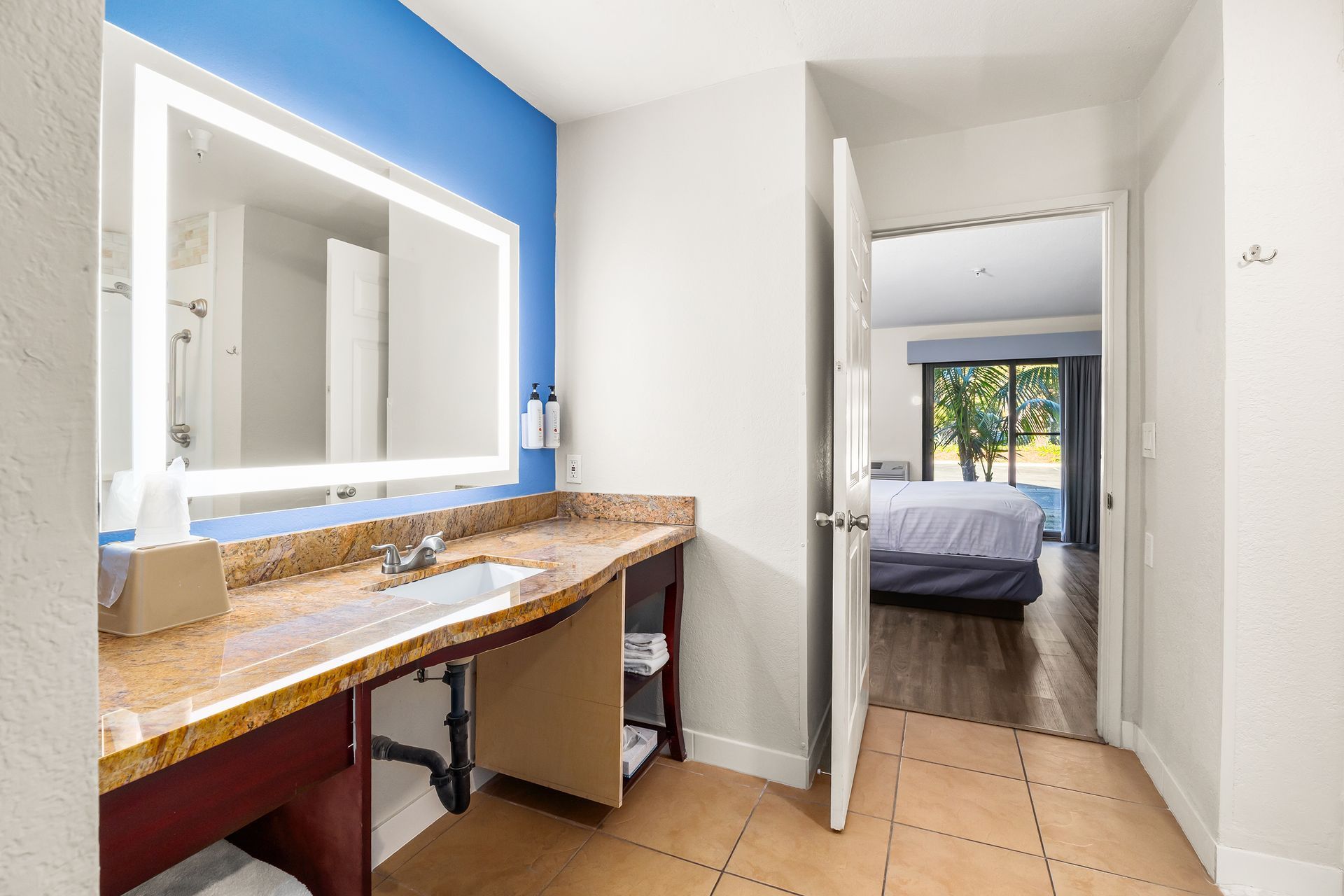 A bathroom vanity with a lighted mirror and granite countertop, seen through a doorway leading to a hotel bedroom.