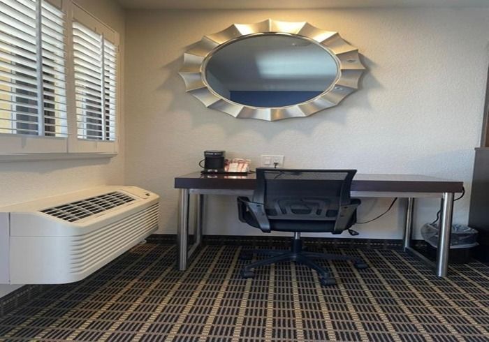 A hotel workspace with a desk, black office chair, and coffee maker, positioned under a large, oval-shaped decorative mirror.
