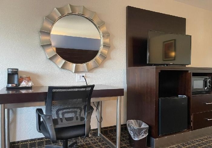 A hotel room desk with an office chair, circular mirror, coffee maker, TV, small refrigerator, and wastebasket.