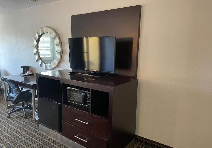 A dark wood media console holds a television and microwave next to a desk, office chair, and circular mirror in a room.