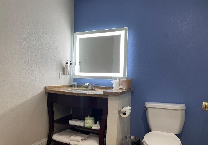 A bathroom vanity with an illuminated mirror against a blue wall, featuring a stone countertop, open shelf, and toilet.