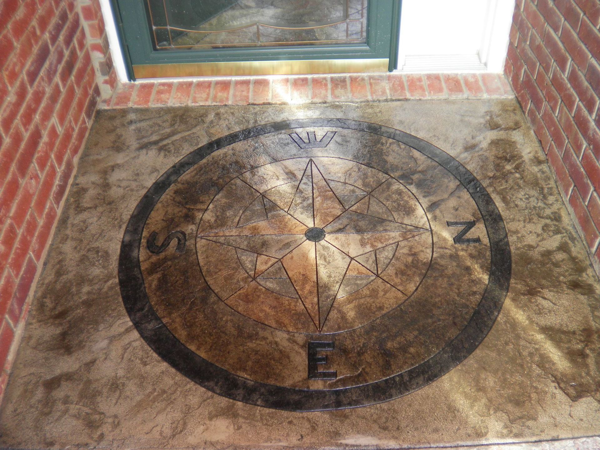 Compass rose design on a concrete entryway, between brick walls and below a green door.