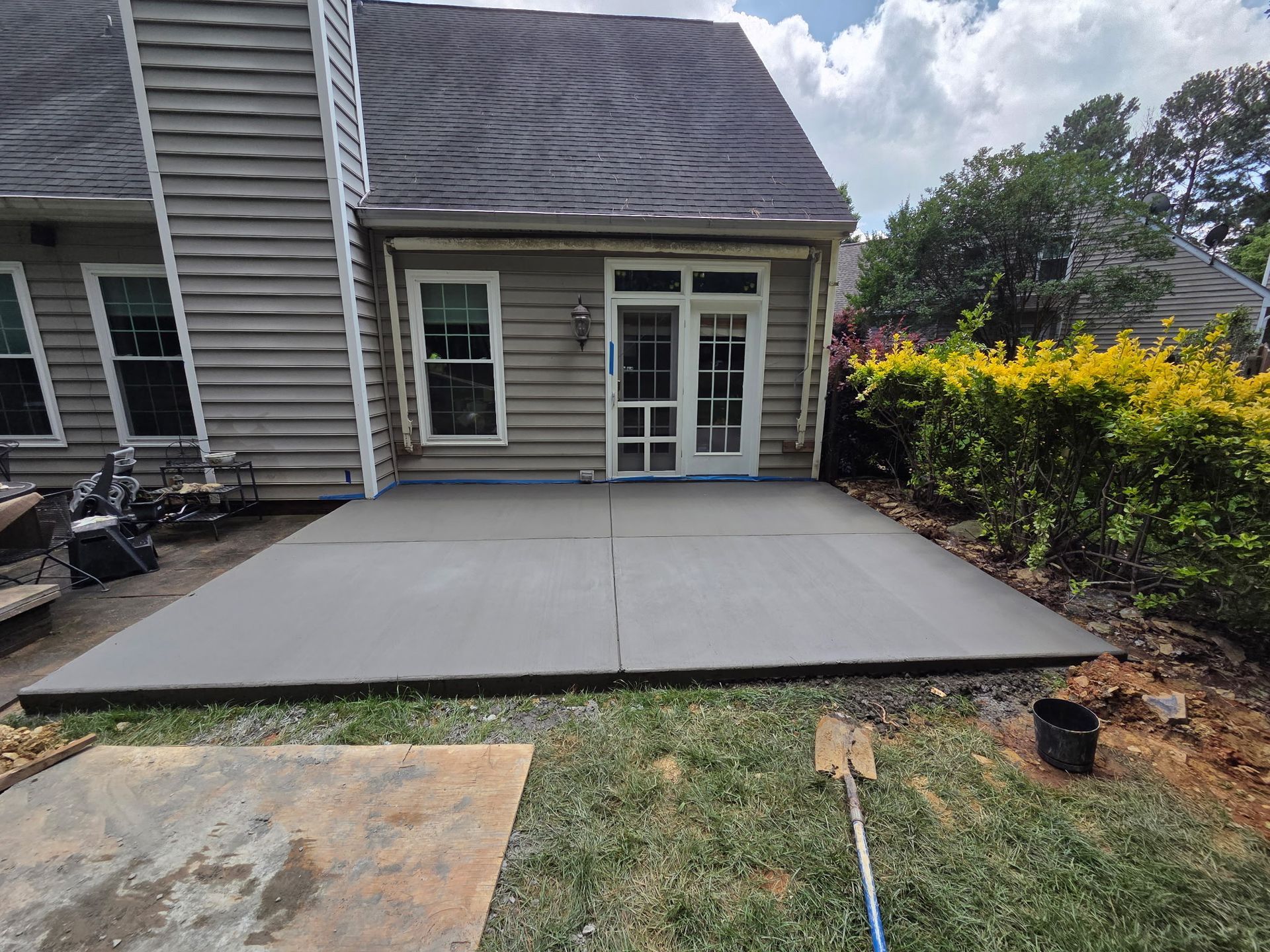A gray concrete patio outside a house with a glass door, surrounded by grass and bushes.