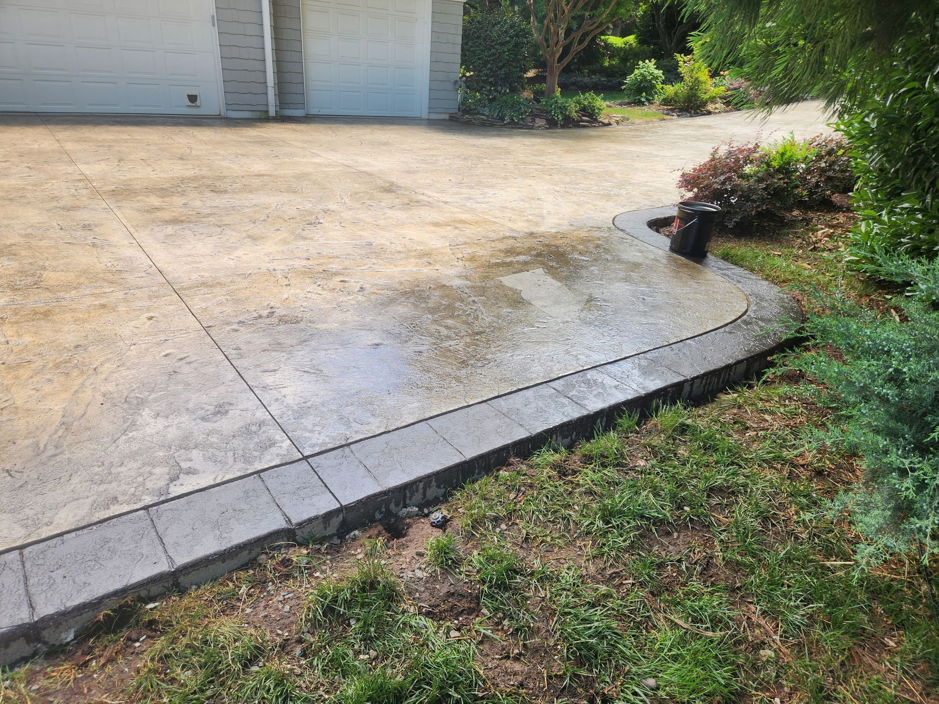 Driveway with brick border, partially wet, near greenery and garage.