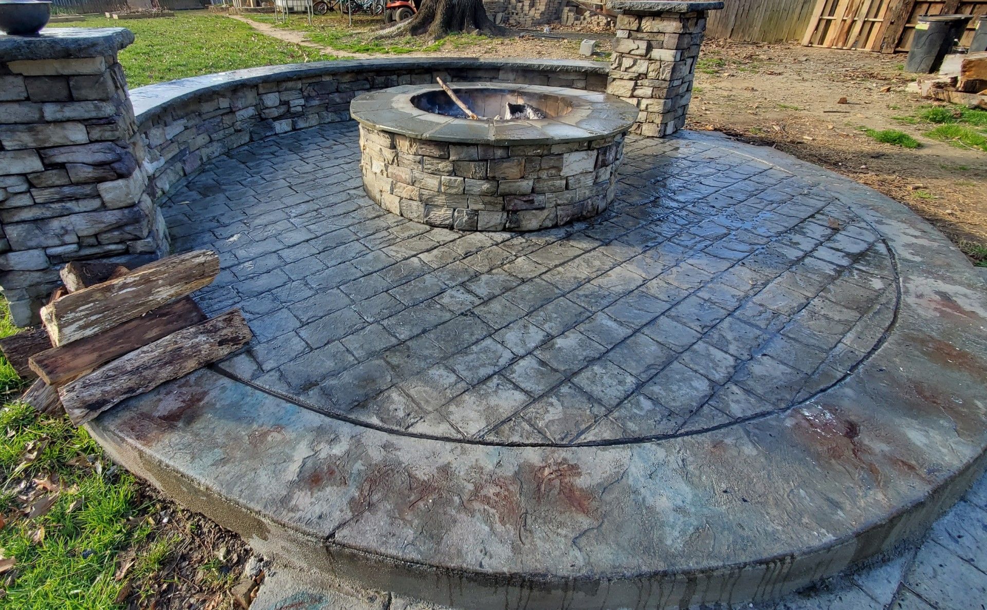 Stone fire pit with surrounding stone patio, posts, and wood logs in a grassy outdoor setting.