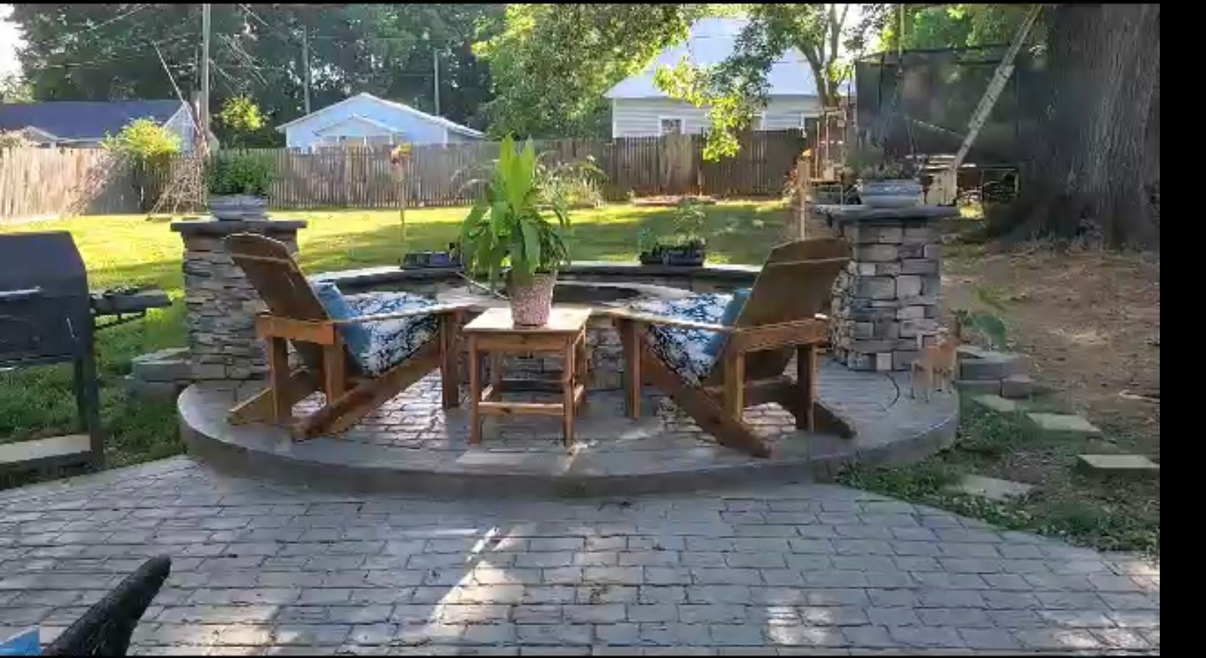 Two wooden chairs, small table with plant, and a fire pit on a stone patio in a backyard.