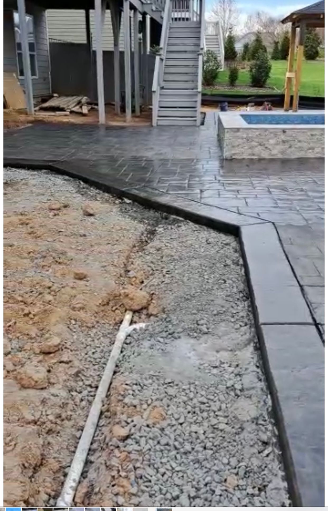 Construction site: brick patio being installed near a deck and small pool. Gravel and dirt visible.