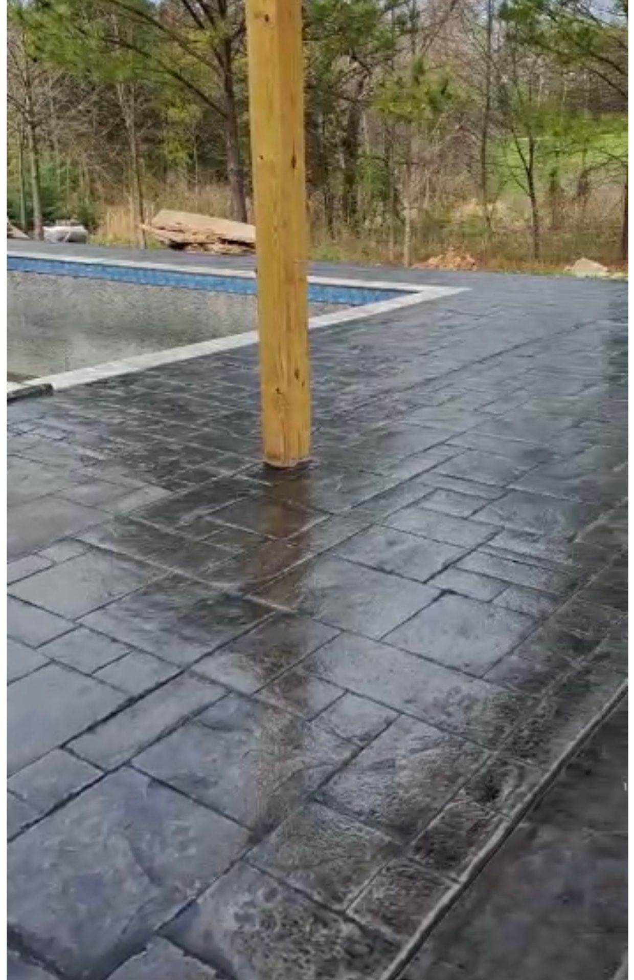 Gray stamped concrete patio with a wooden post, next to a pool in a wooded area.