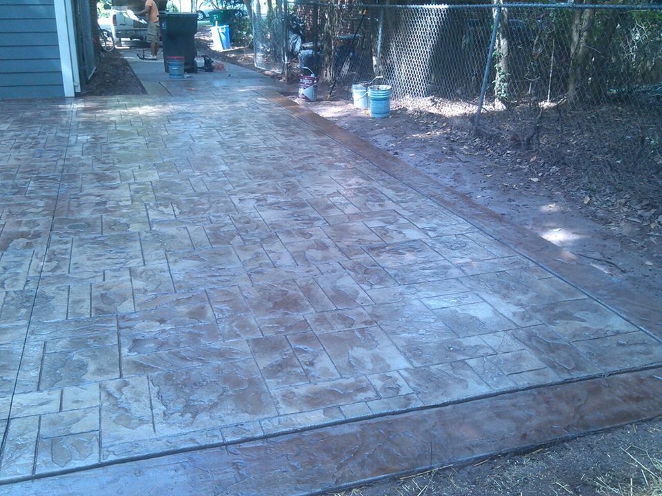 Stamped concrete patio with brown brick pattern, next to a chain-link fence and trees.