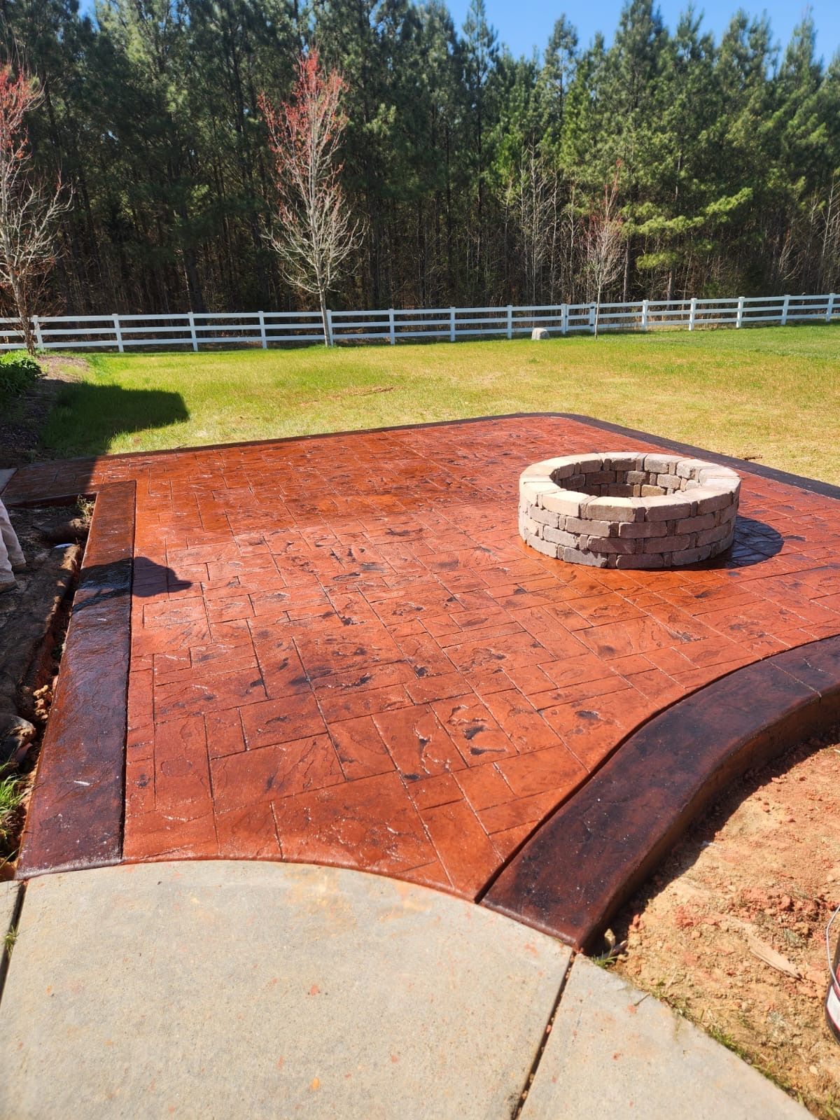 Stamped concrete patio with fire pit, surrounded by grass and trees; white fence in background.