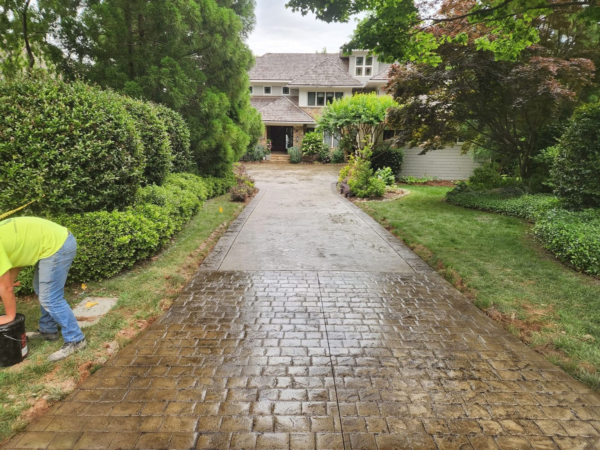 Person working on a wet, patterned brick driveway leading to a house. Green bushes border the drive.