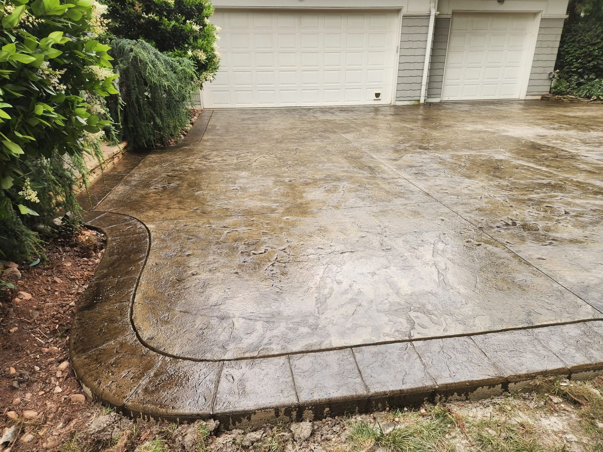 Wet concrete driveway with decorative border, next to garage and greenery.