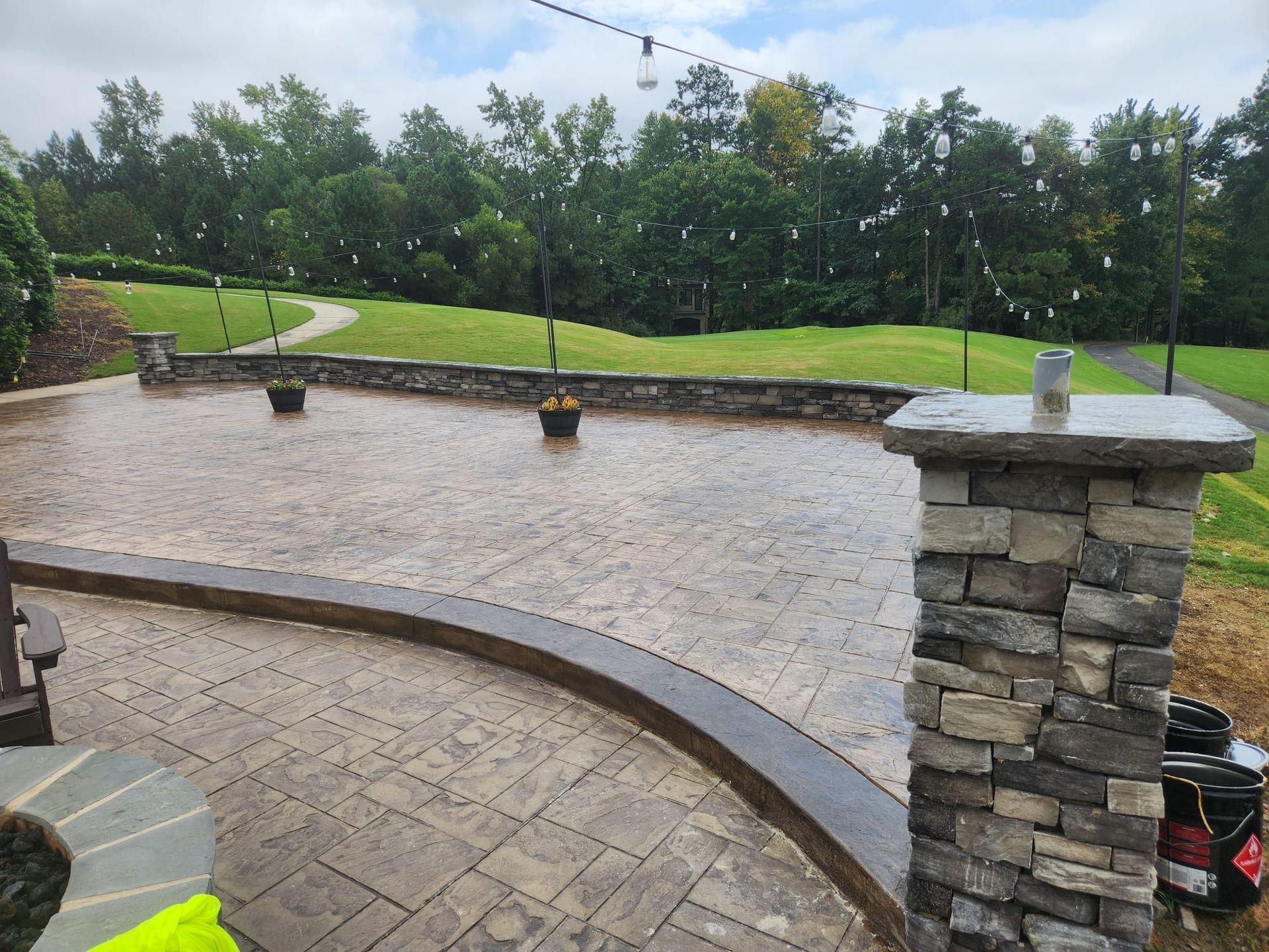 Wet stone patio with stone pillars, in front of grass and trees. Cloudy day.