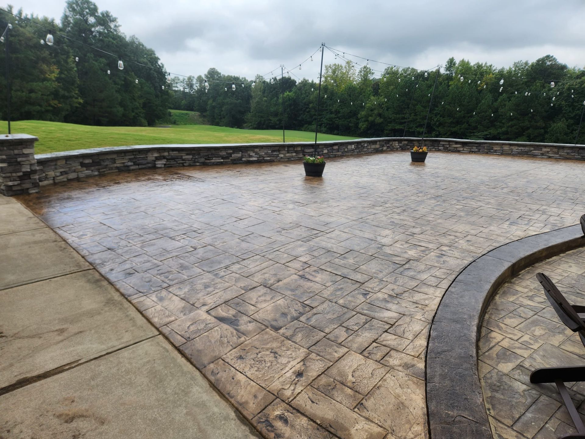Stamped concrete patio with brick border, overlooking a grassy lawn and trees under a cloudy sky.