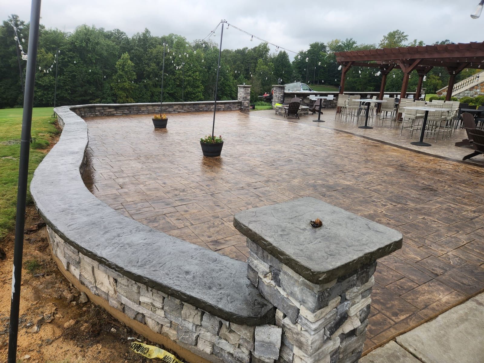 Stone patio with a low wall, tables, and a pergola, set in an outdoor area.