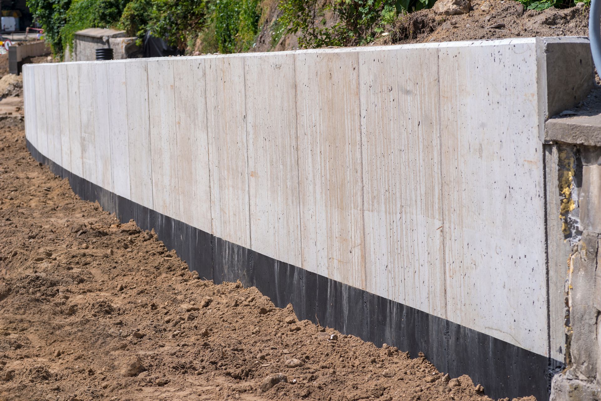 Concrete retaining wall, light gray with dark band at base, set in brown soil.