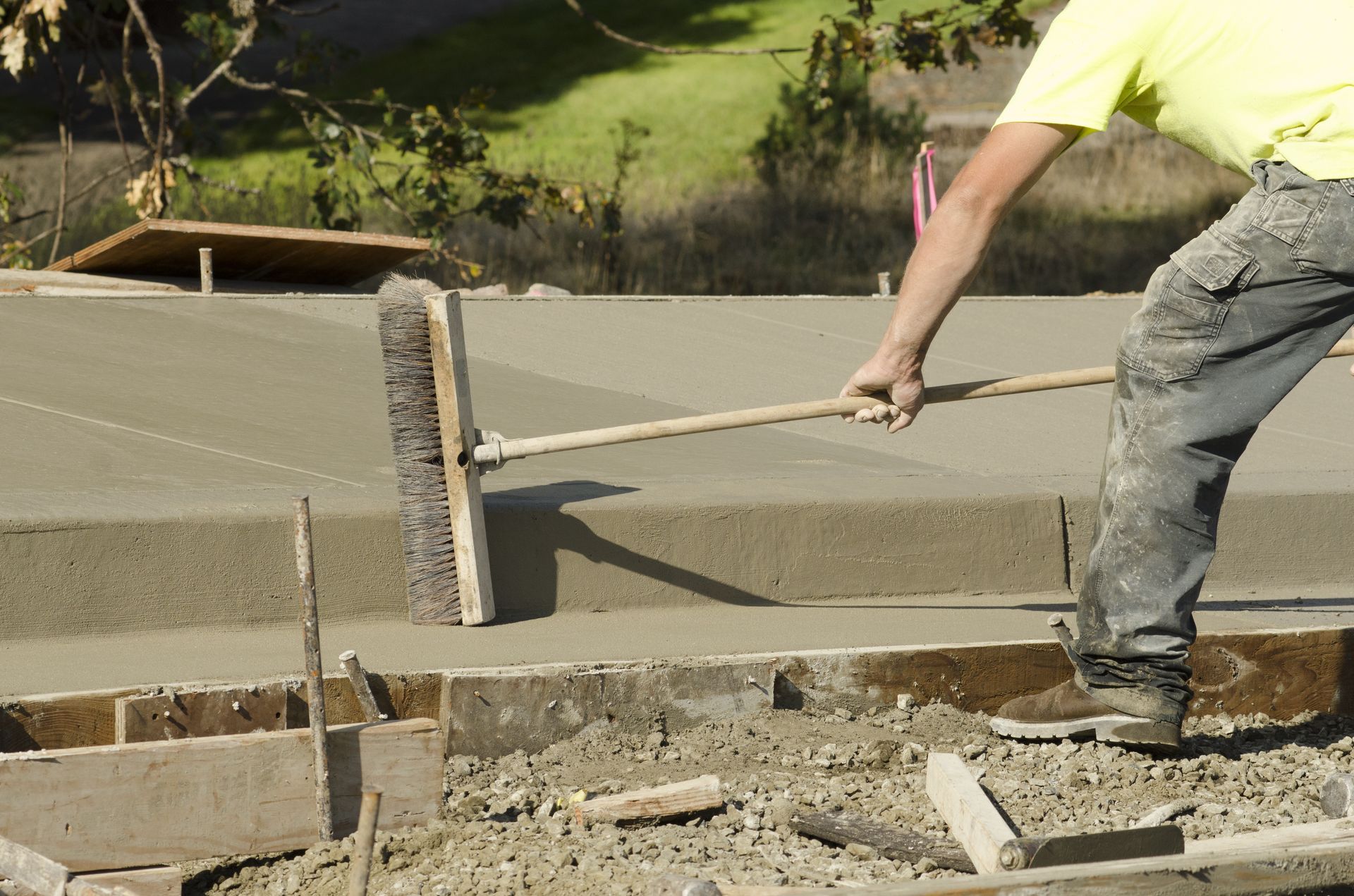 Construction worker brushing wet concrete with a long-handled broom, outdoors.