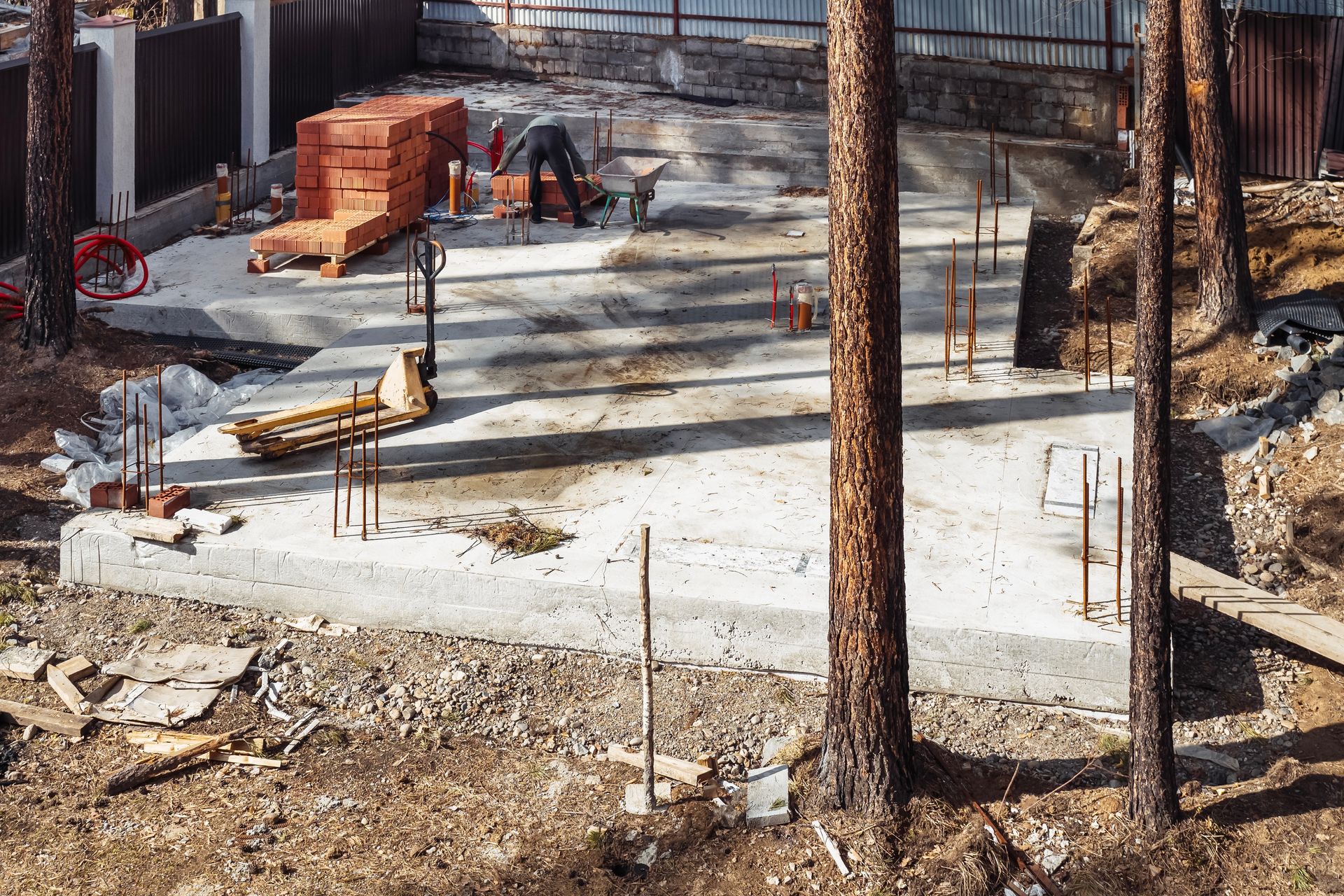 Construction site: concrete foundation, bricks, rebar, surrounded by trees. Workers visible.