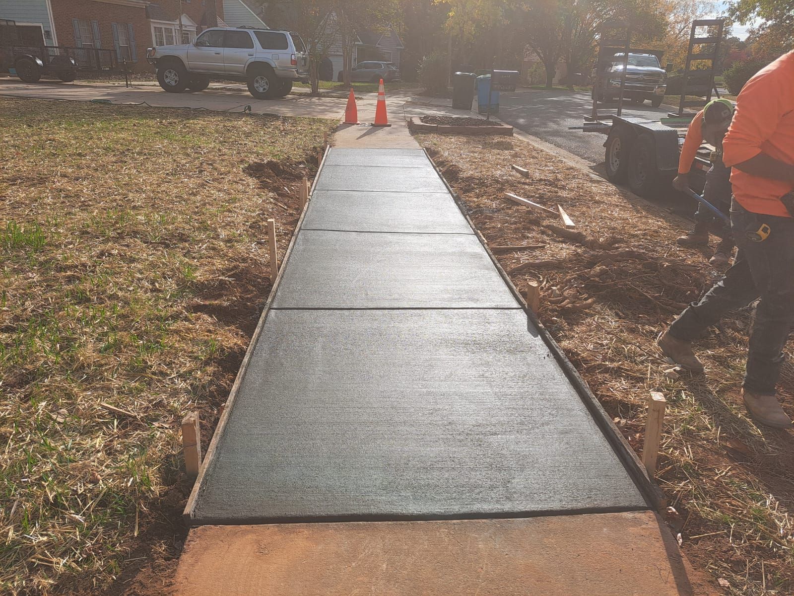 Newly paved concrete sidewalk. Worker in orange jacket uses a blower, beside grass and a truck.