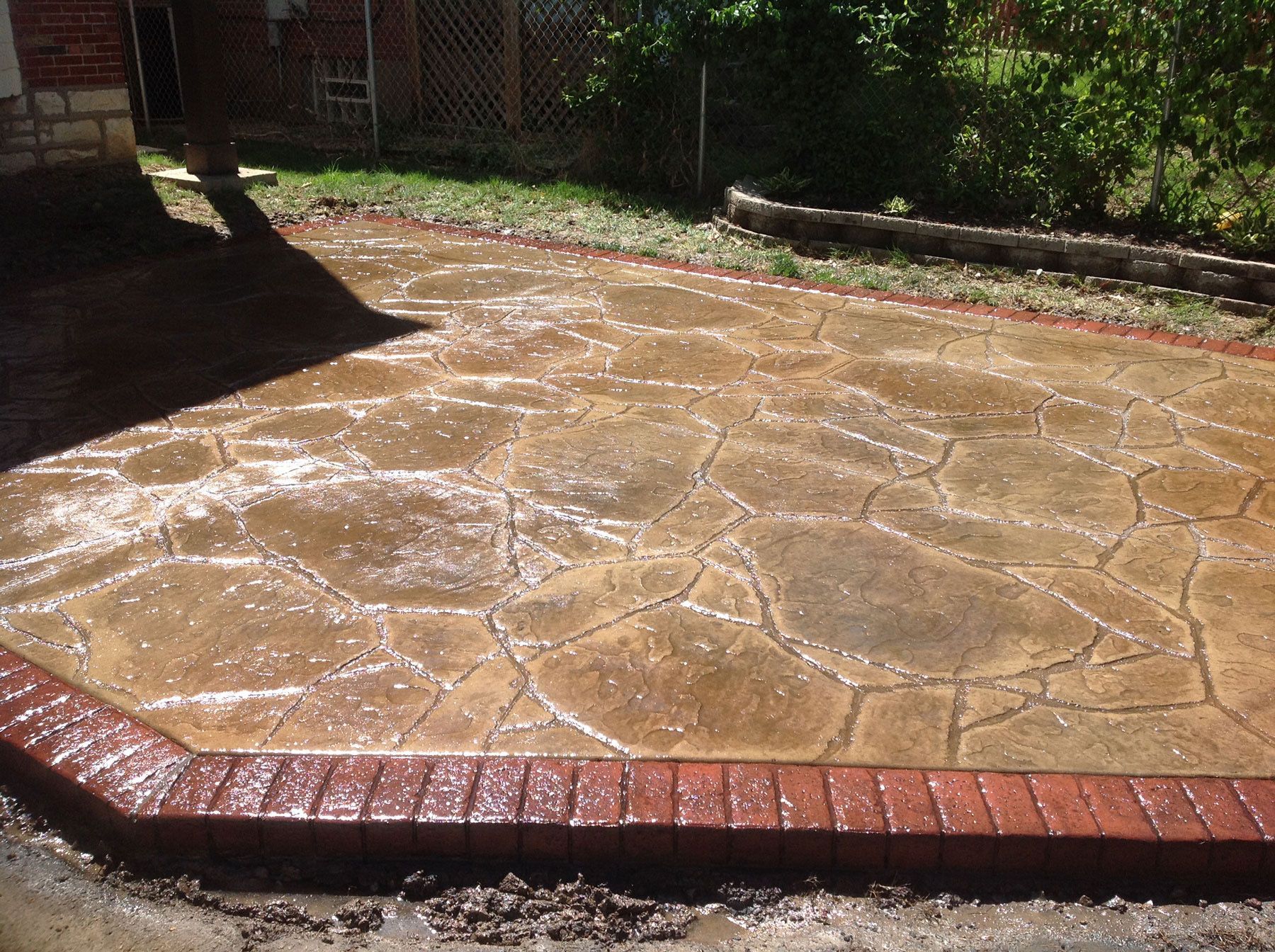 Brown and red patterned concrete patio with a brick border, beside grass.