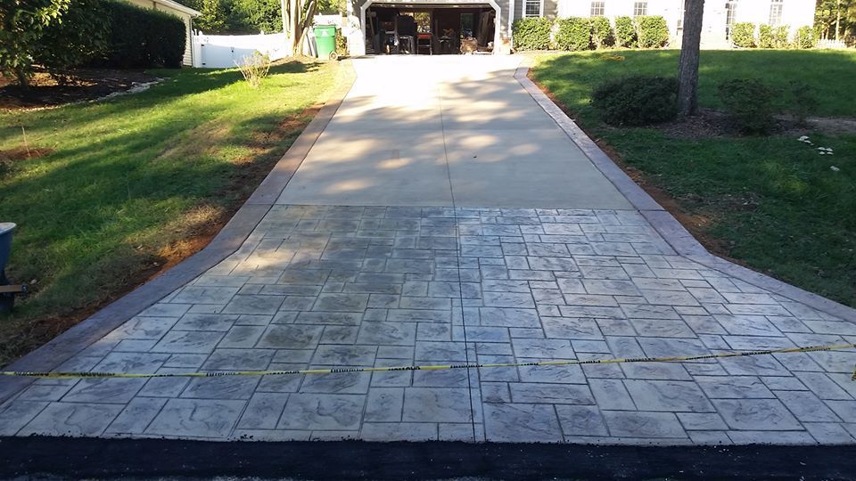 Concrete driveway with stamped stone pattern leading to a garage, bordered by grass and trees.