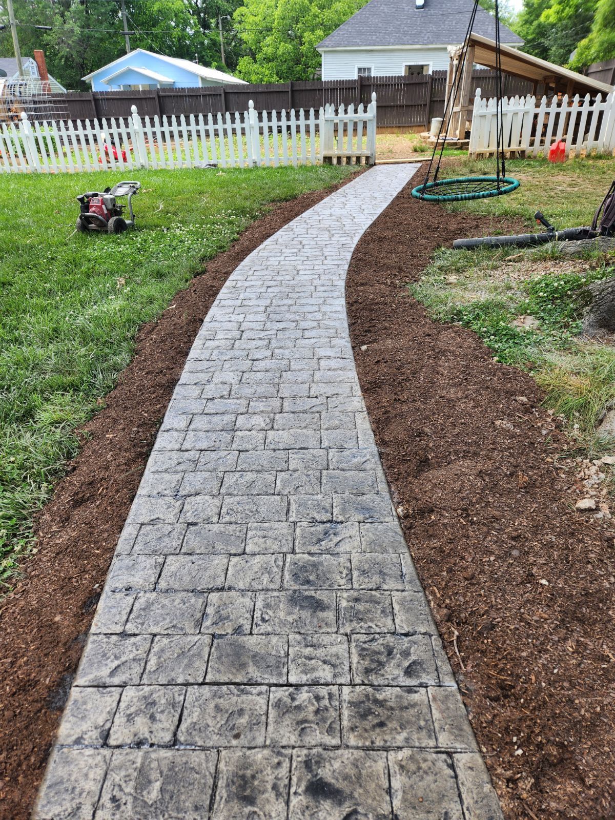 Concrete path, textured to look like brick, winds through a yard. Mulch borders the path.
